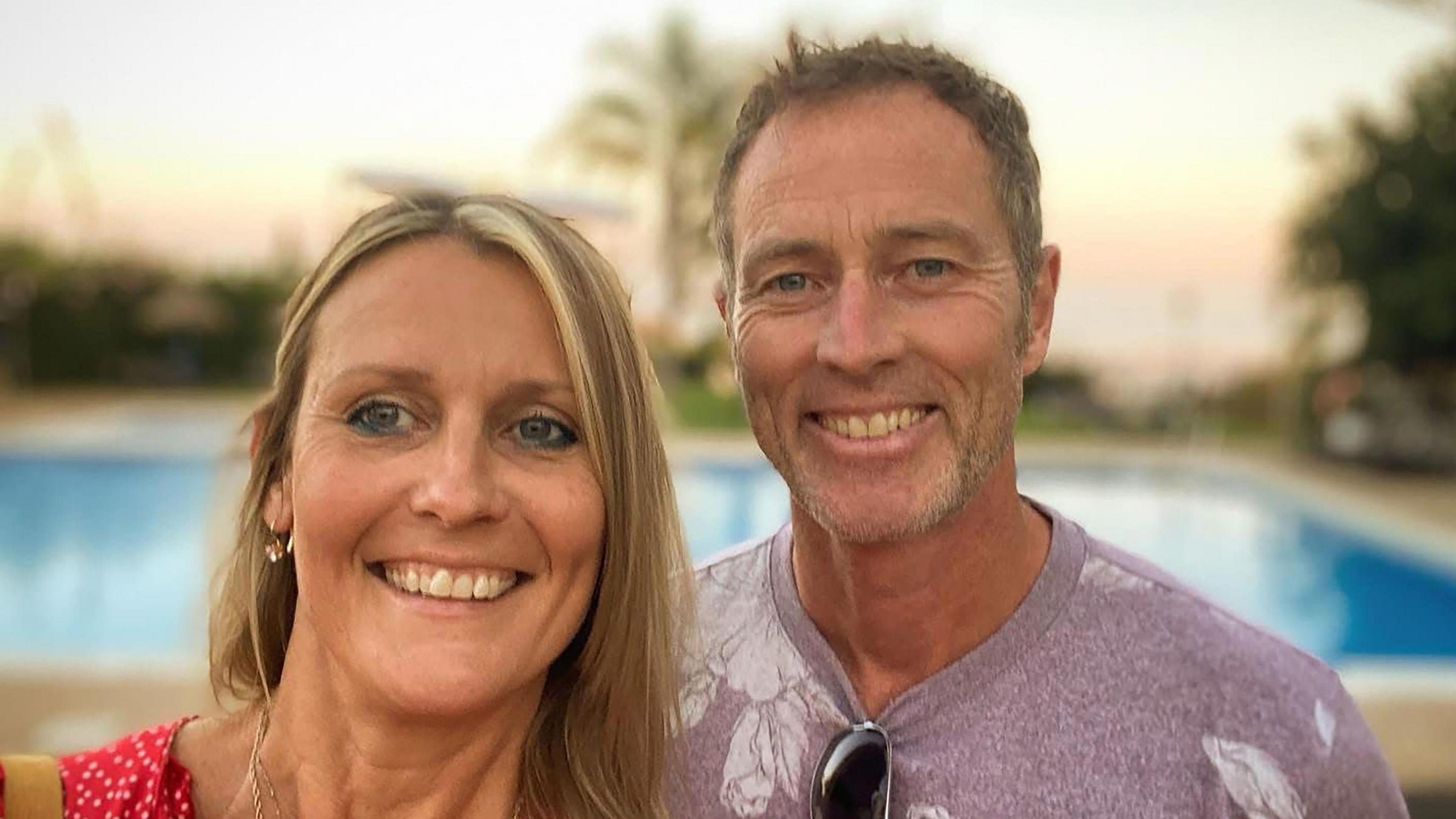 A man and woman smile on holiday. The woman has shoulder length blonde hair and is wearing a red top, the man has short brown hair, stubble and a lilac t-shirt. There is a pool behind them.