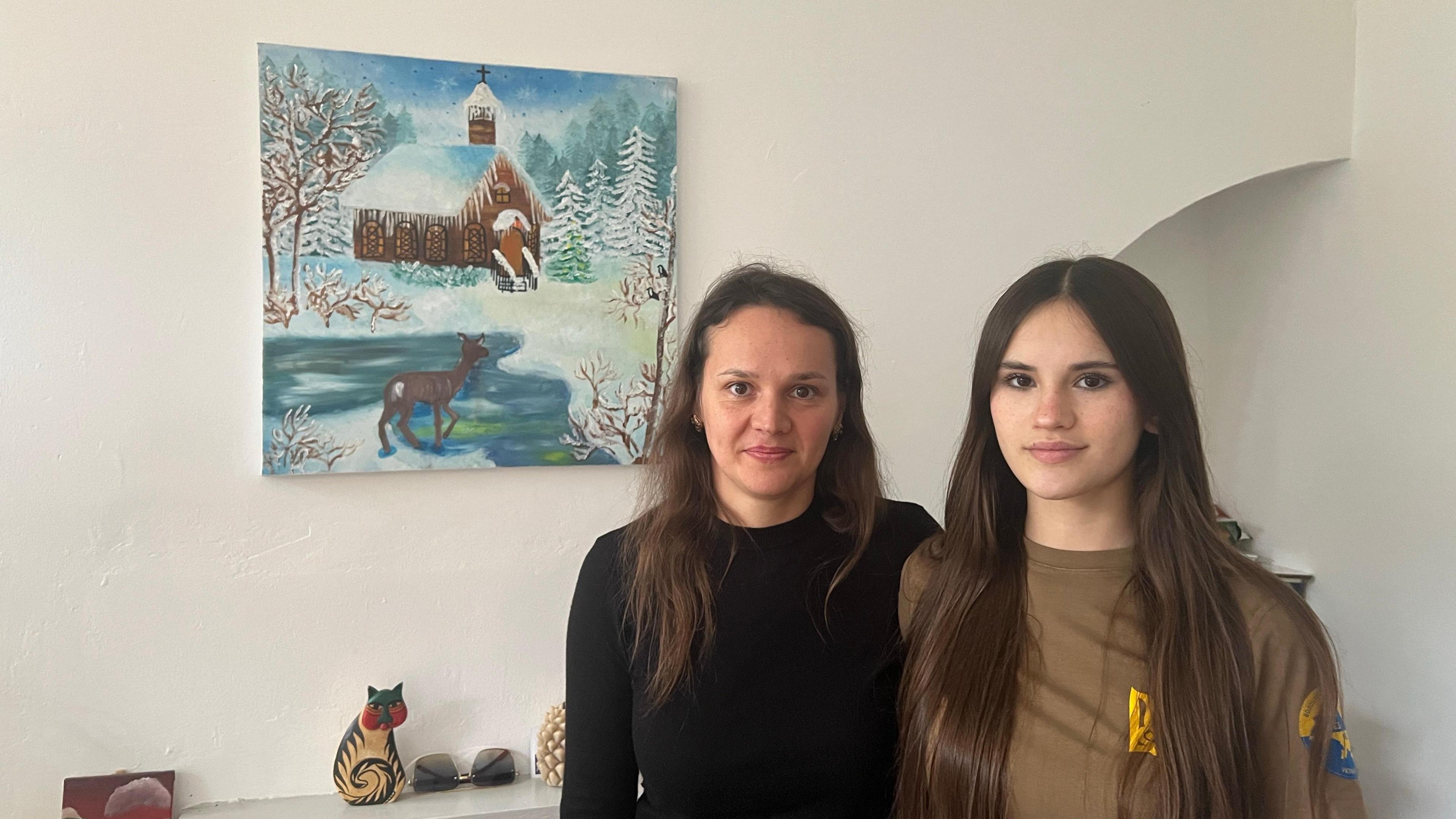 A woman with brown hair dressed all in black is stood next to a girl who is wearing a brown T-shirt. They are standing in front of a painting of a wintery scene depicting a cottage in the snow with a deer next to a lake.