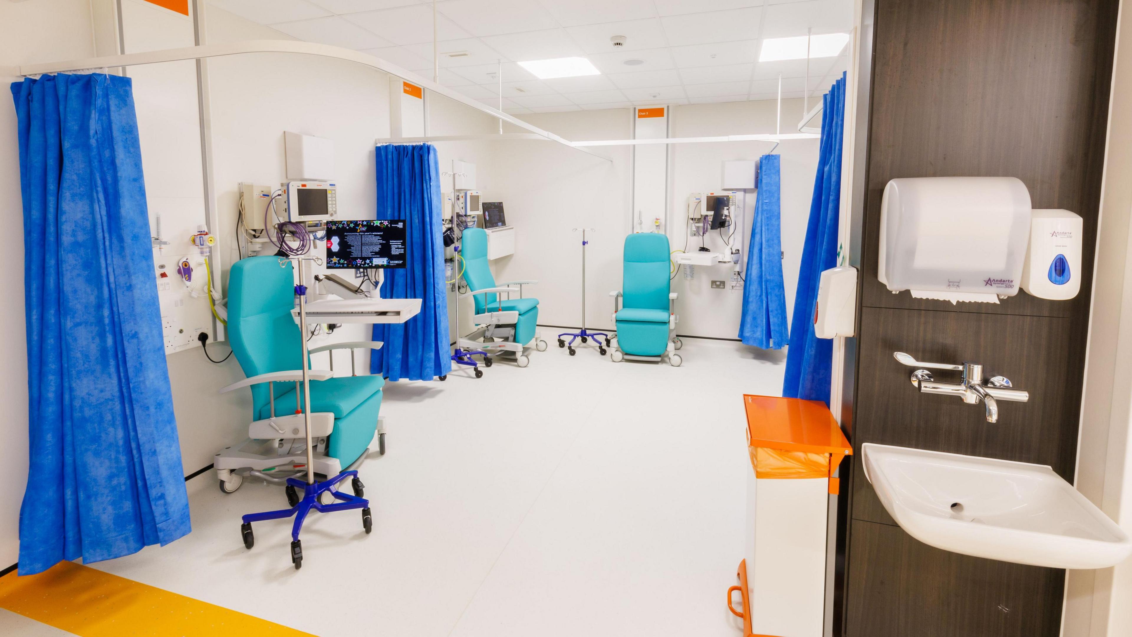 A clean-looking medical room with blue paper curtains and three blue recliner chairs in it. In the foreground, outside the ward, is a sink attached to the wall with soap and paper towels.