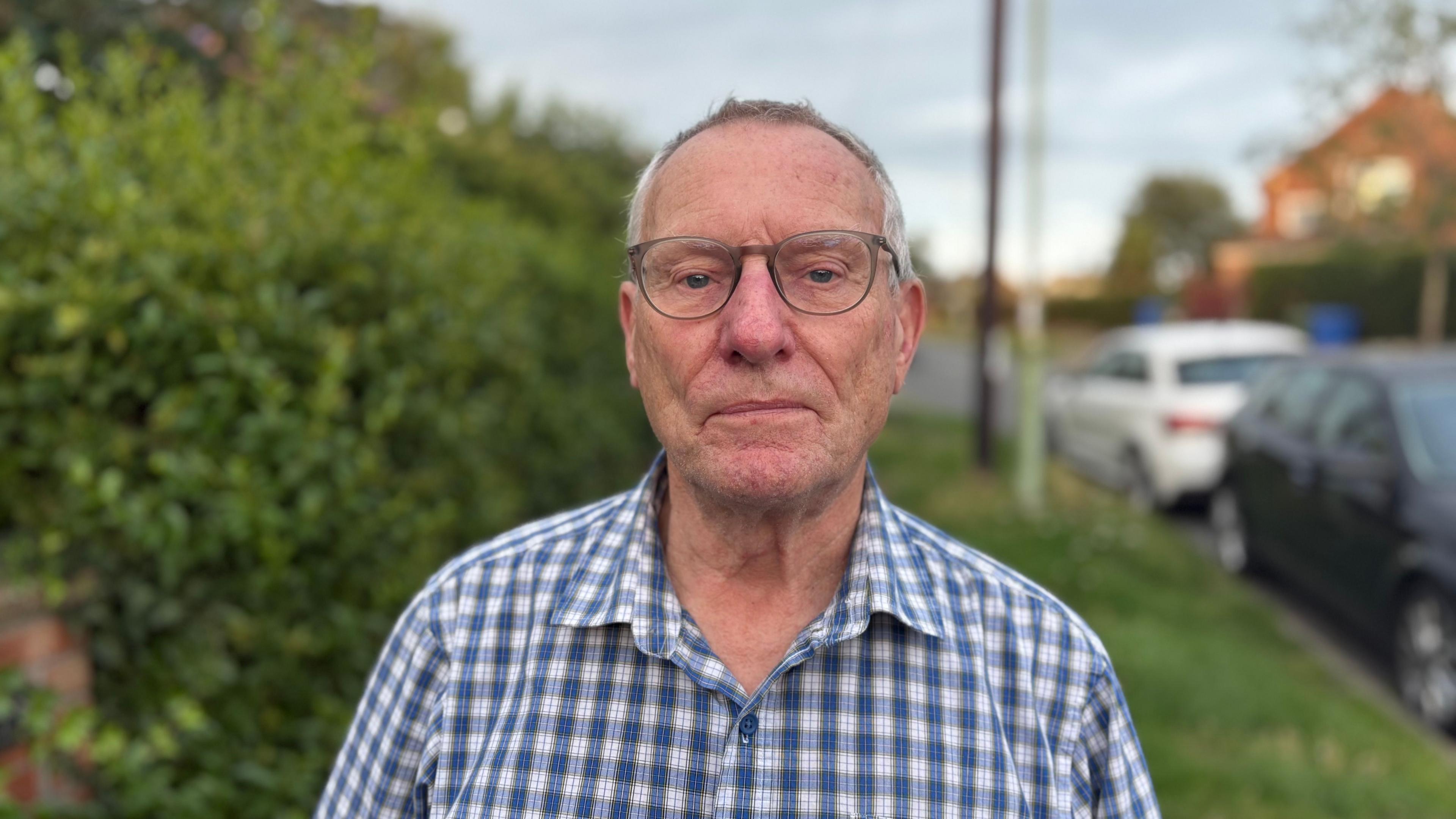 Peter Byatt smiles at the camera. He has short grey hair, has glasses and wears a chequered blue and white shirt.