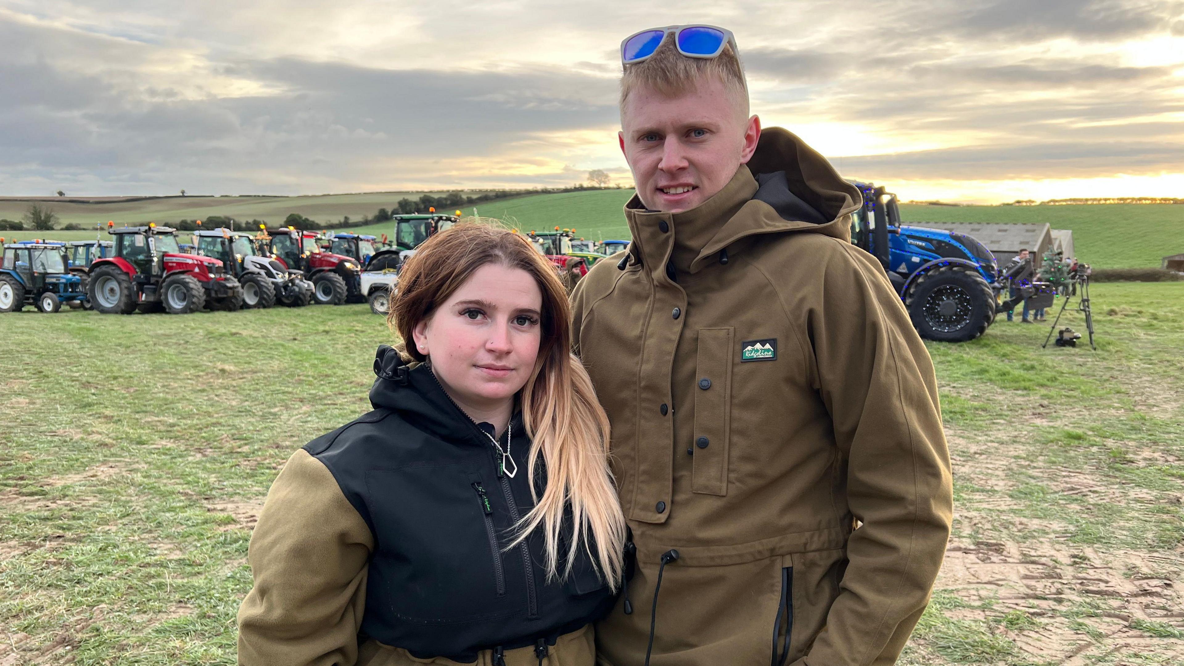 Bryony is wearing a black and green coat. She has brown hair with blond highlights. Kian is wearing a green coat and he has blond hair. The couple are standing in a field with rows of tractors behind them.