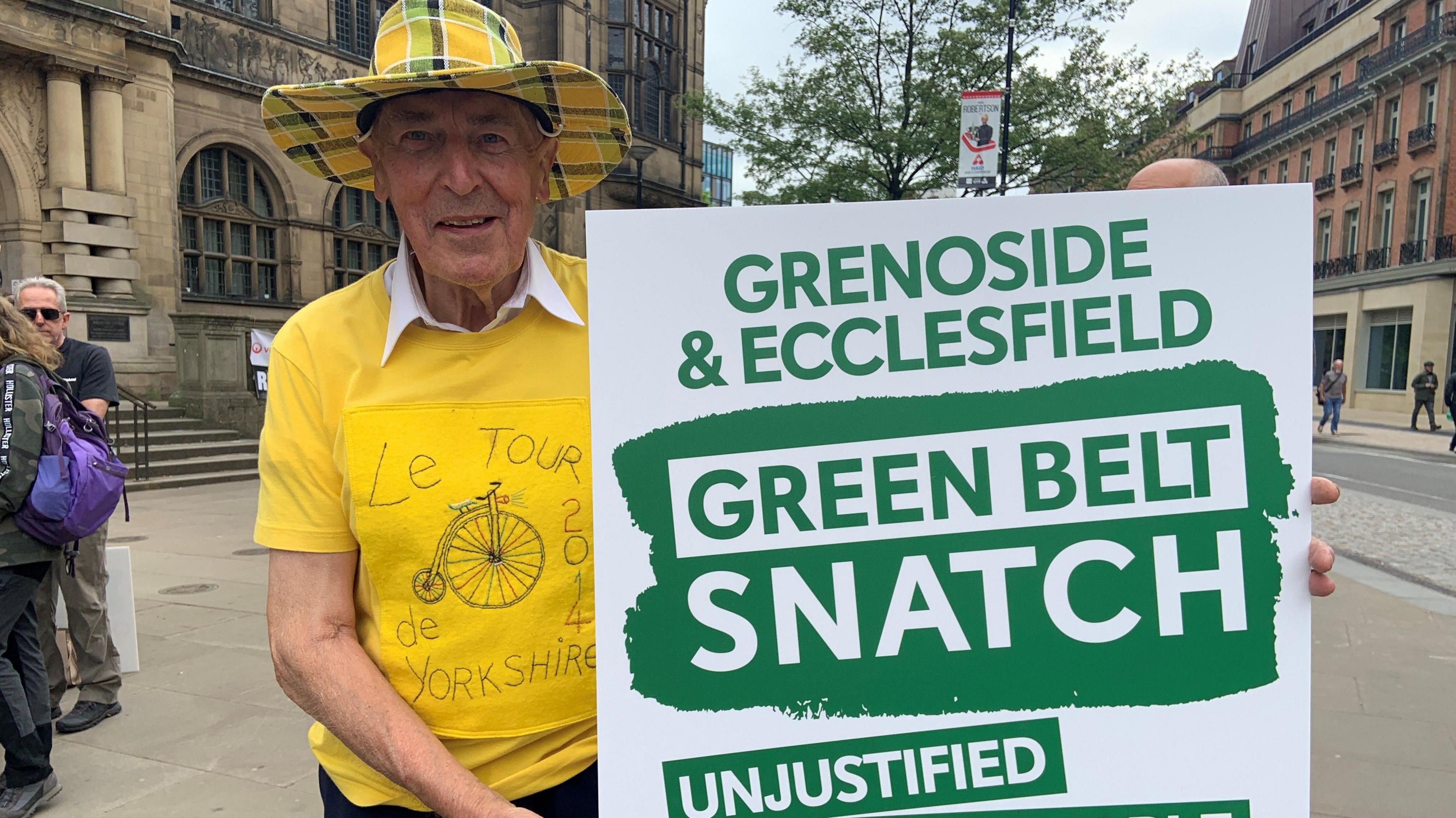 An elderly man wearing a yellow check hat and a yellow t-shirt with the words Le Tour de Yorkshire written on it. He is holding a green-and-white placard.