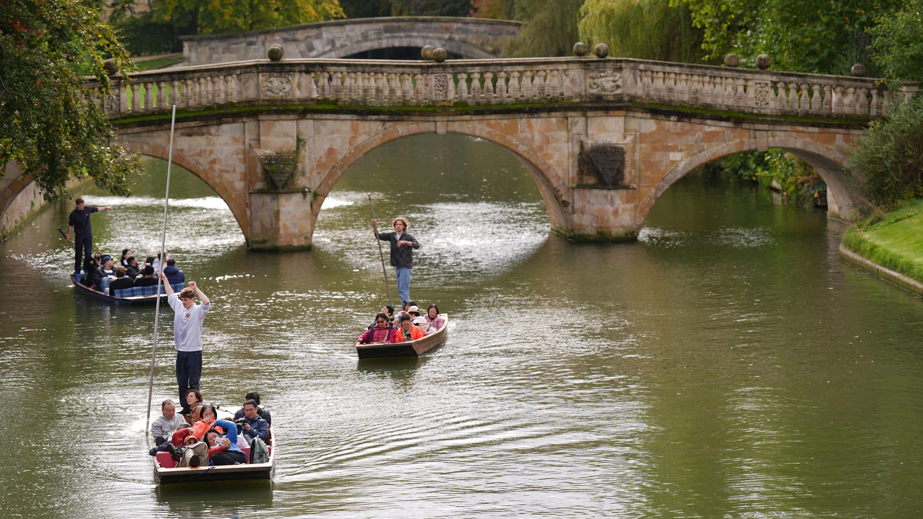 A picture of punting on the River Cam in Cambridge in 2024, with three punts containing tourists. In the background are two bridges. It is a sunny day.