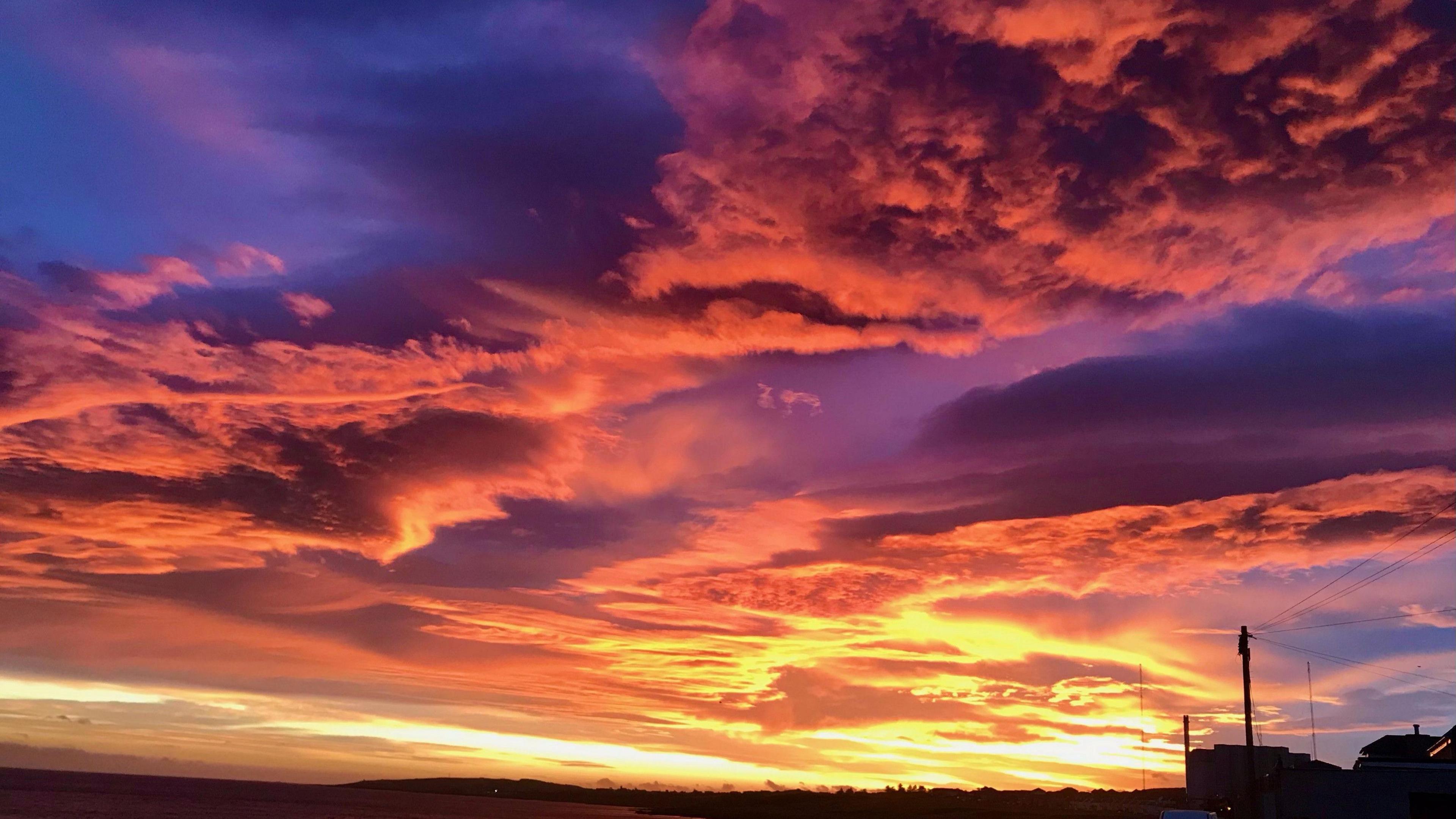 The image captures a sunset over a body of water with a shoreline in the foreground. The sky is vibrant hues of orange, pink, and purple. The clouds are thick and textured, adding depth and dimension to the scene. In the distance, a silhouette of a coastal town is visible.