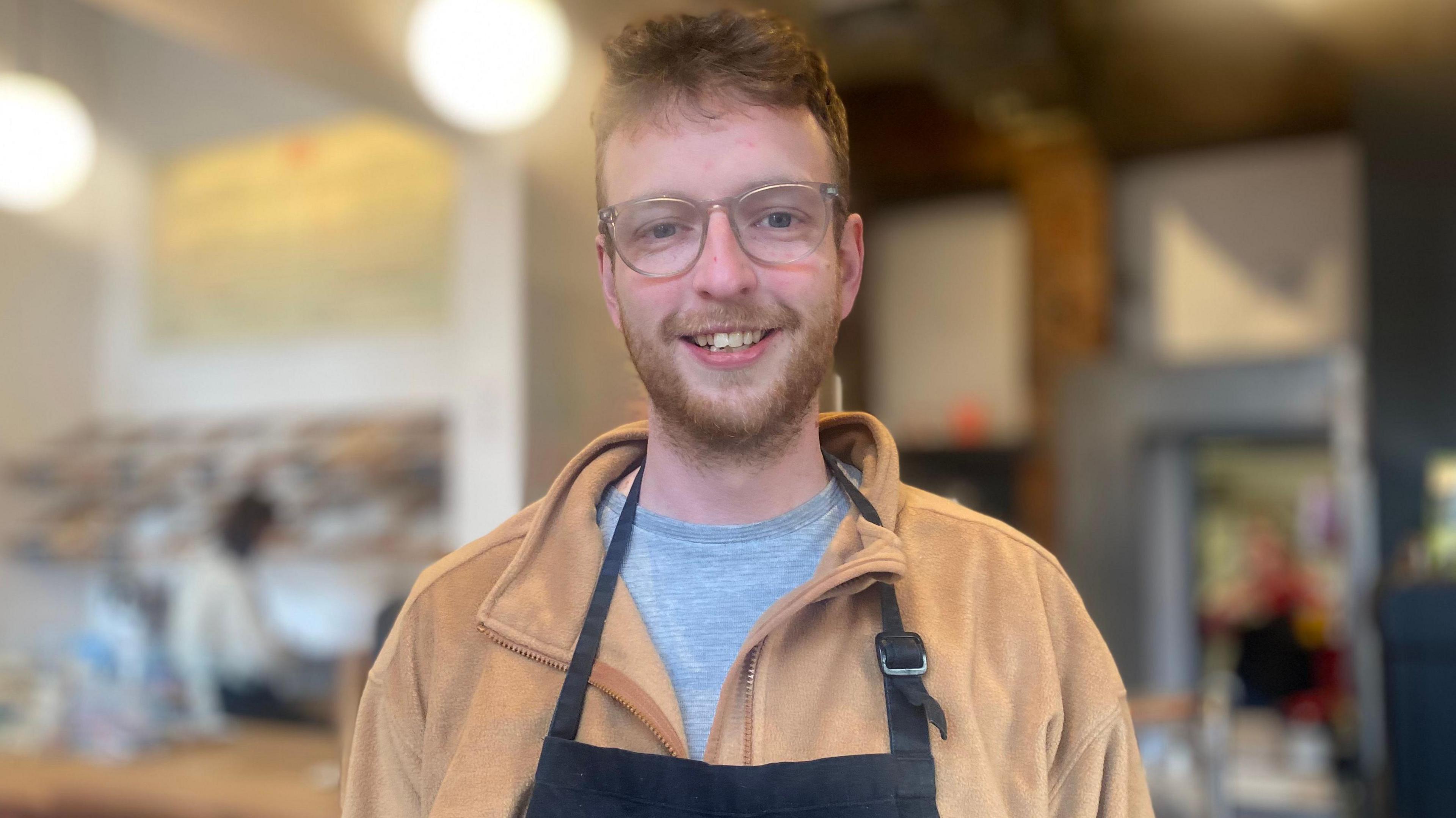 Steven smiles at the camera. He is wearing a brown fleece, grey t-shirt and black apron. He has brown hair and wars glasses. Behind him is the shop counter.