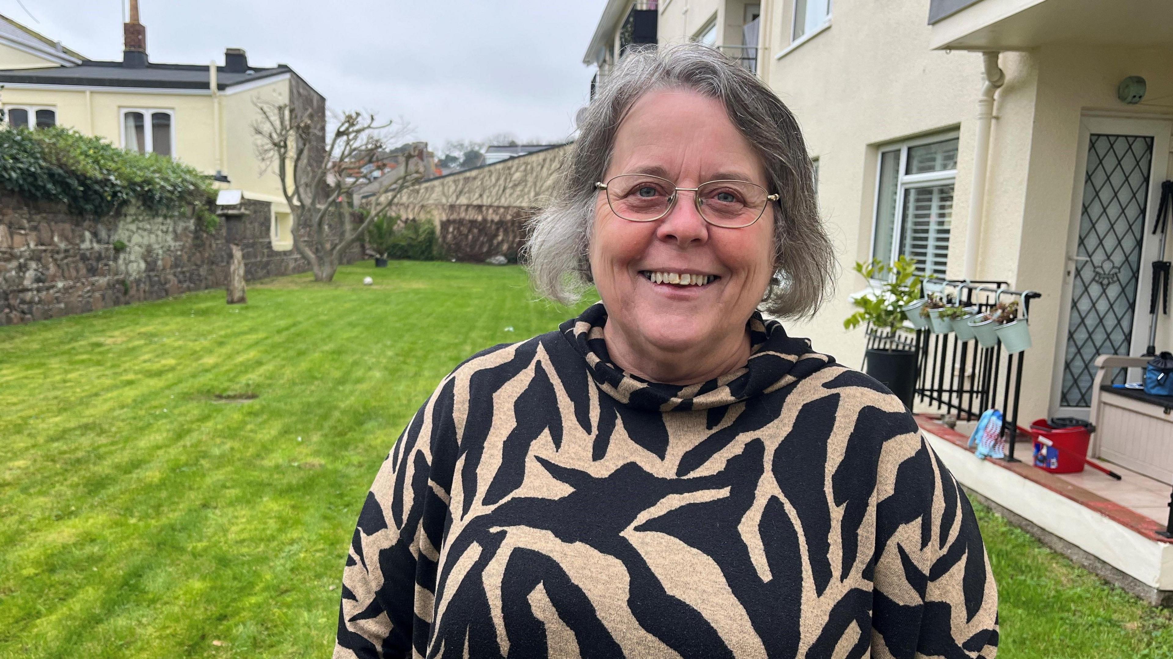 Jeannine Carey standing in a garden with a white building in the background. She is looking into the camera, while wearing an animal print top. She has short grey hair and glasses. 