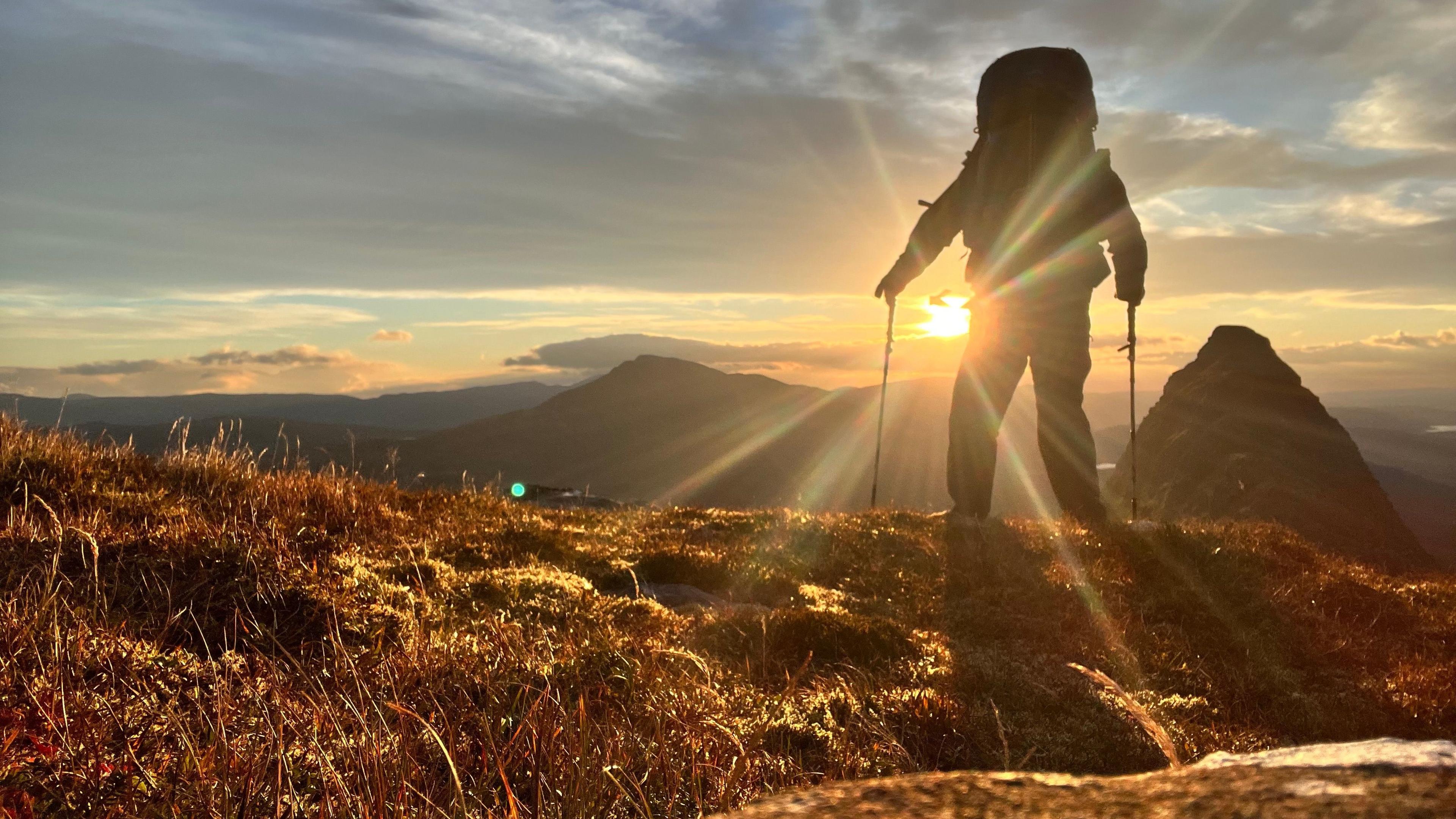 A person standing on a grassy hilltop, equipped with hiking poles, gazing toward the sunrise. The low sun casts long shadows, turning the hiker into a striking silhouette. In the background, distant mountains stretch across the horizon beneath a partly cloudy sky, all bathed in a warm, golden light that enhances the dramatic and peaceful atmosphere.