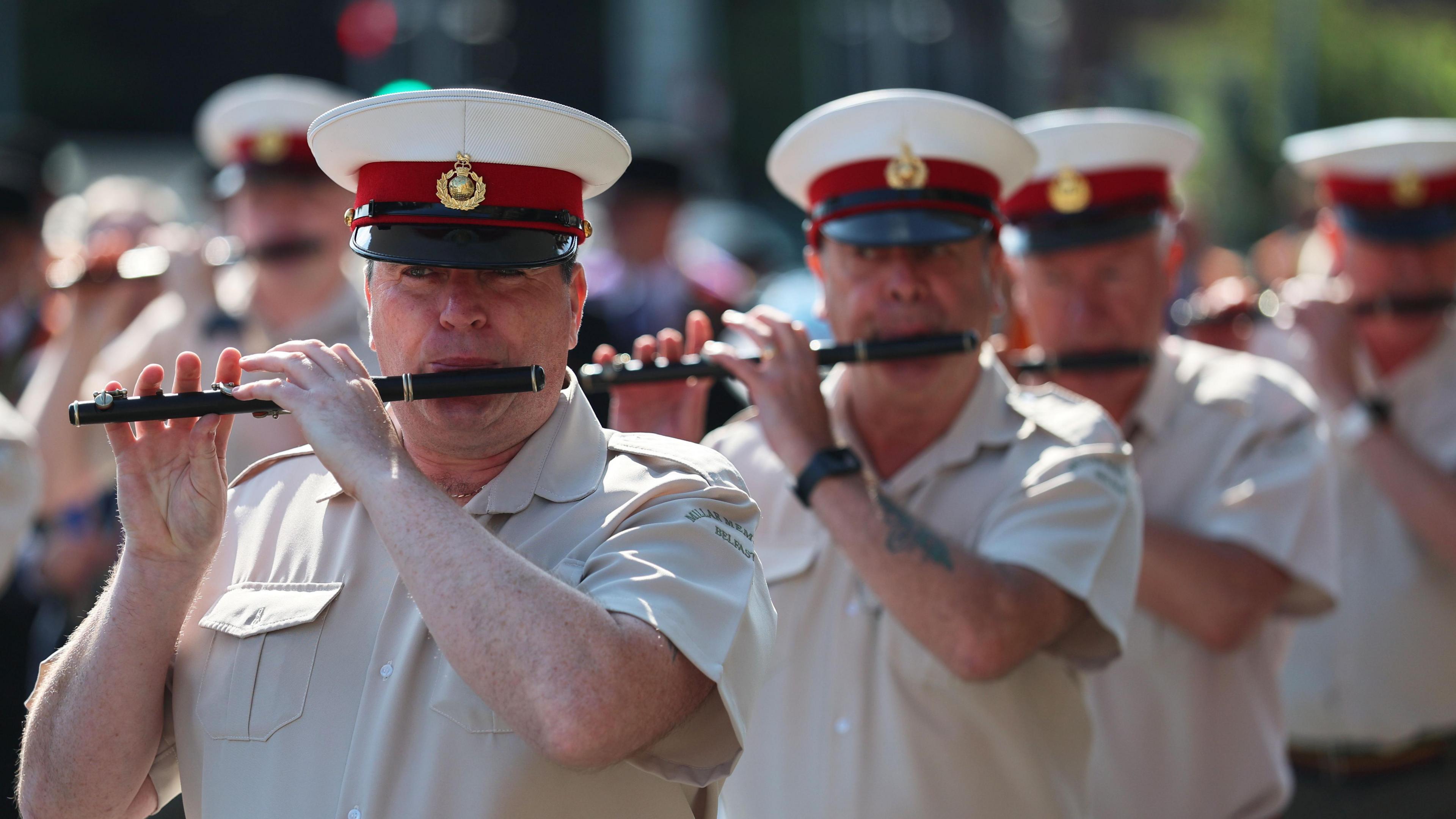 A number of men are walking and playing the flute. It is a sunny day and they are wearing short sleeved shirts and hats which have a white top, black peak and a maroon band with an insignia on the front. The focus is on the man in the front - the others following behind become increasingly blurred.