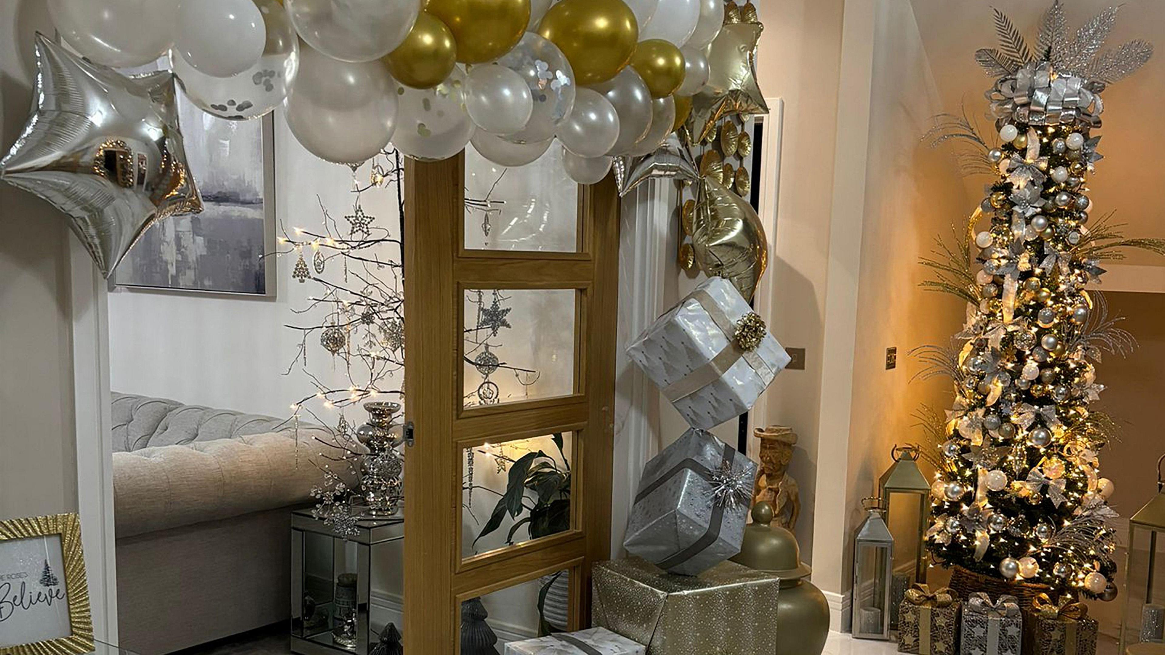 A Christmas themed hallway. Gold and white balloons and a stack of gold and white presents border a living room door. In the corner is a tall Christmas tree decorated with white and gold Christmas baubles and lit up in warm white fairy lights. 