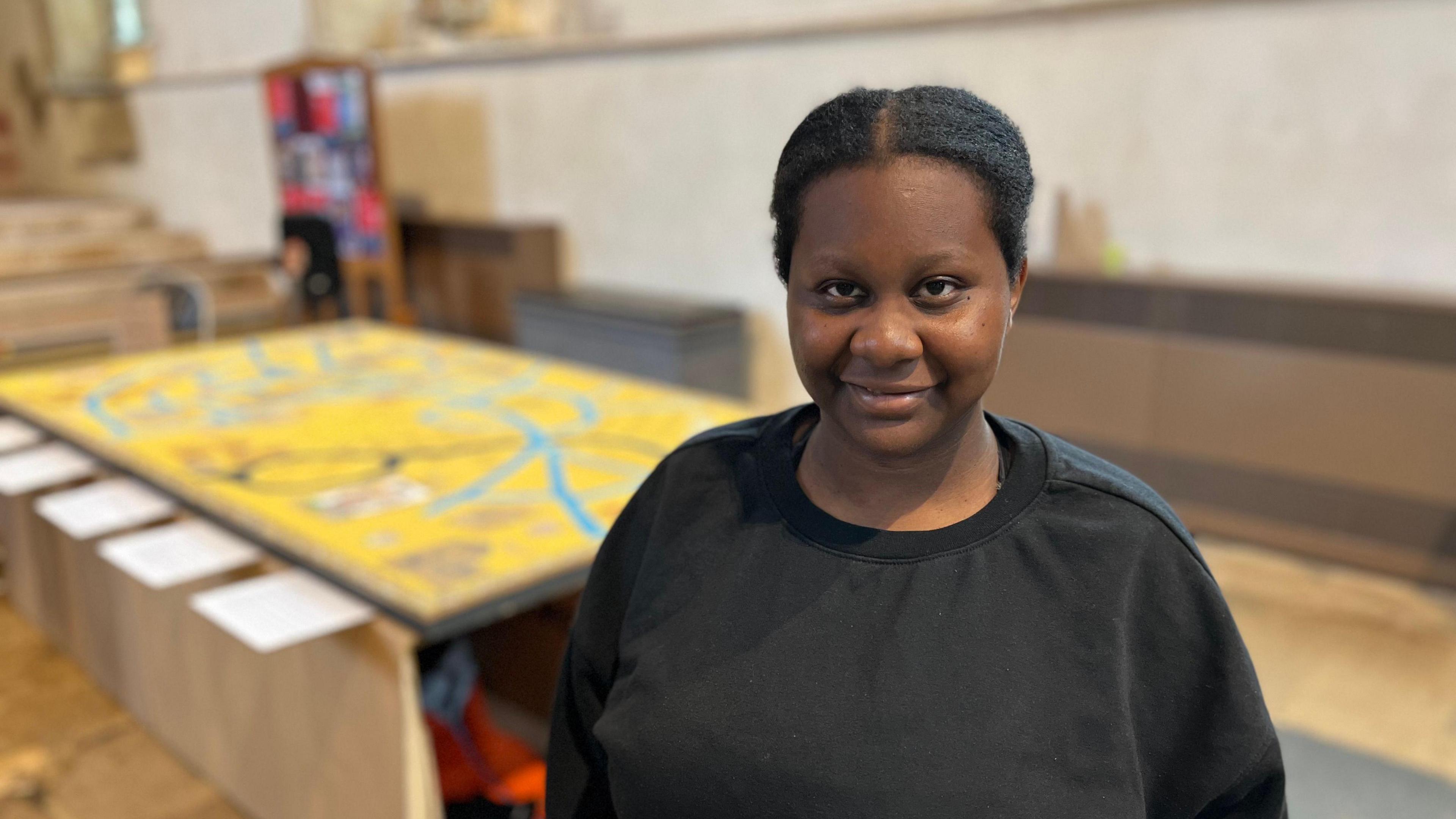 Janet Mboulota wearing a black crew neck sweatshirt. In the background is the artwork, out of focus.