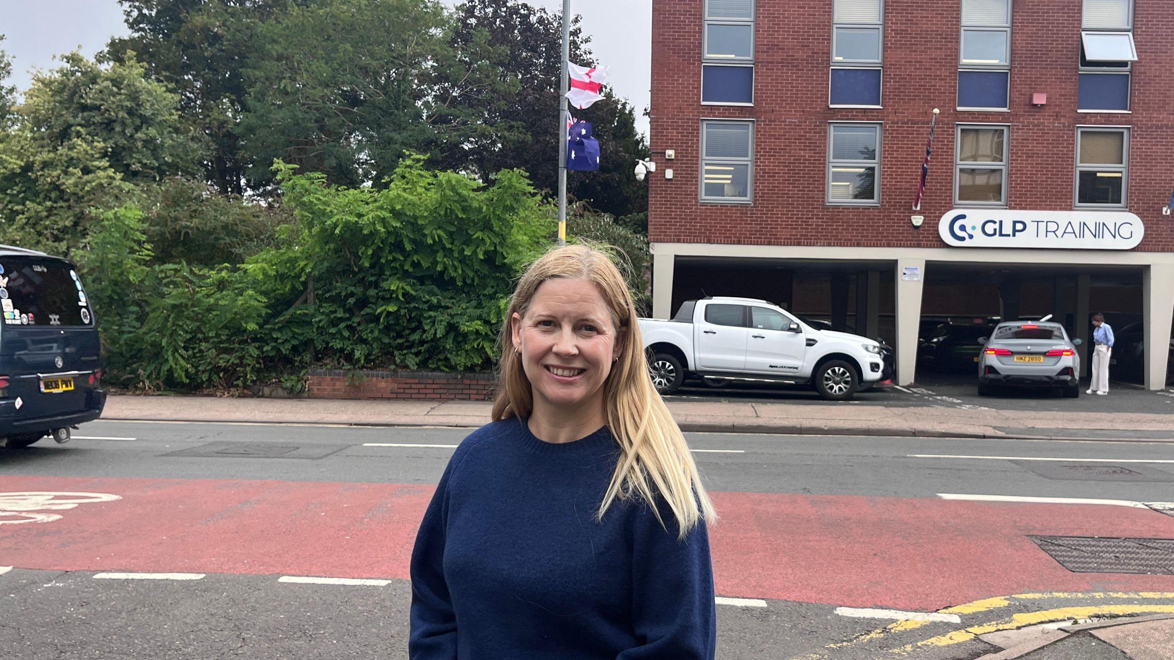 A blonde wearing a blue crew-necked jumper stood alongside a road with a lamppost in the background with an England and an Australian flag flying