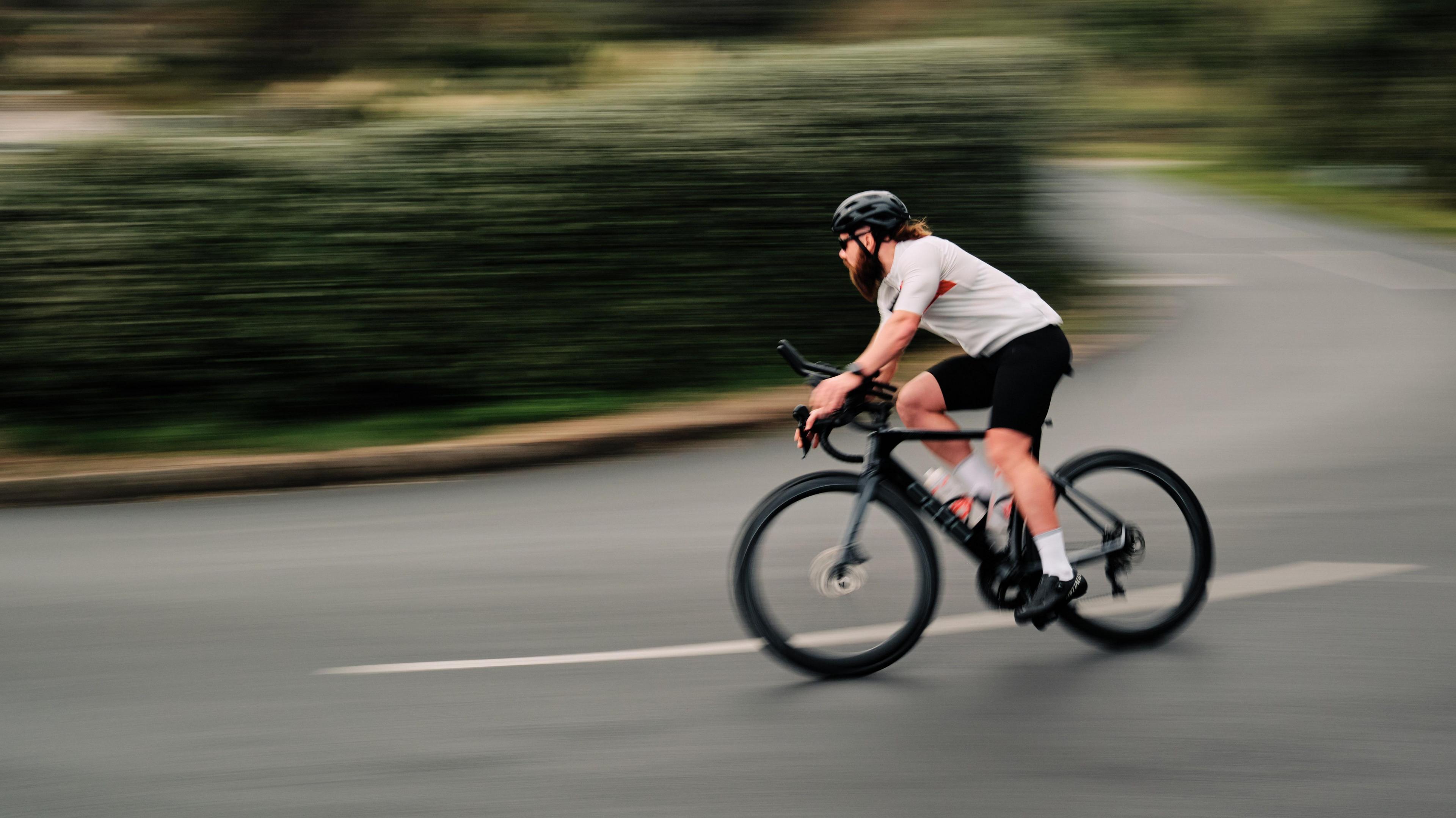 Joe leans forward as he cycles along. He's wearing a black helmet and sunglasses with a white short-sleeved top with black shorts. His ponytail sticks out from under the helmet as he cycles around a bend in the road and the background is blured behind him as Joe is cycling at speed.