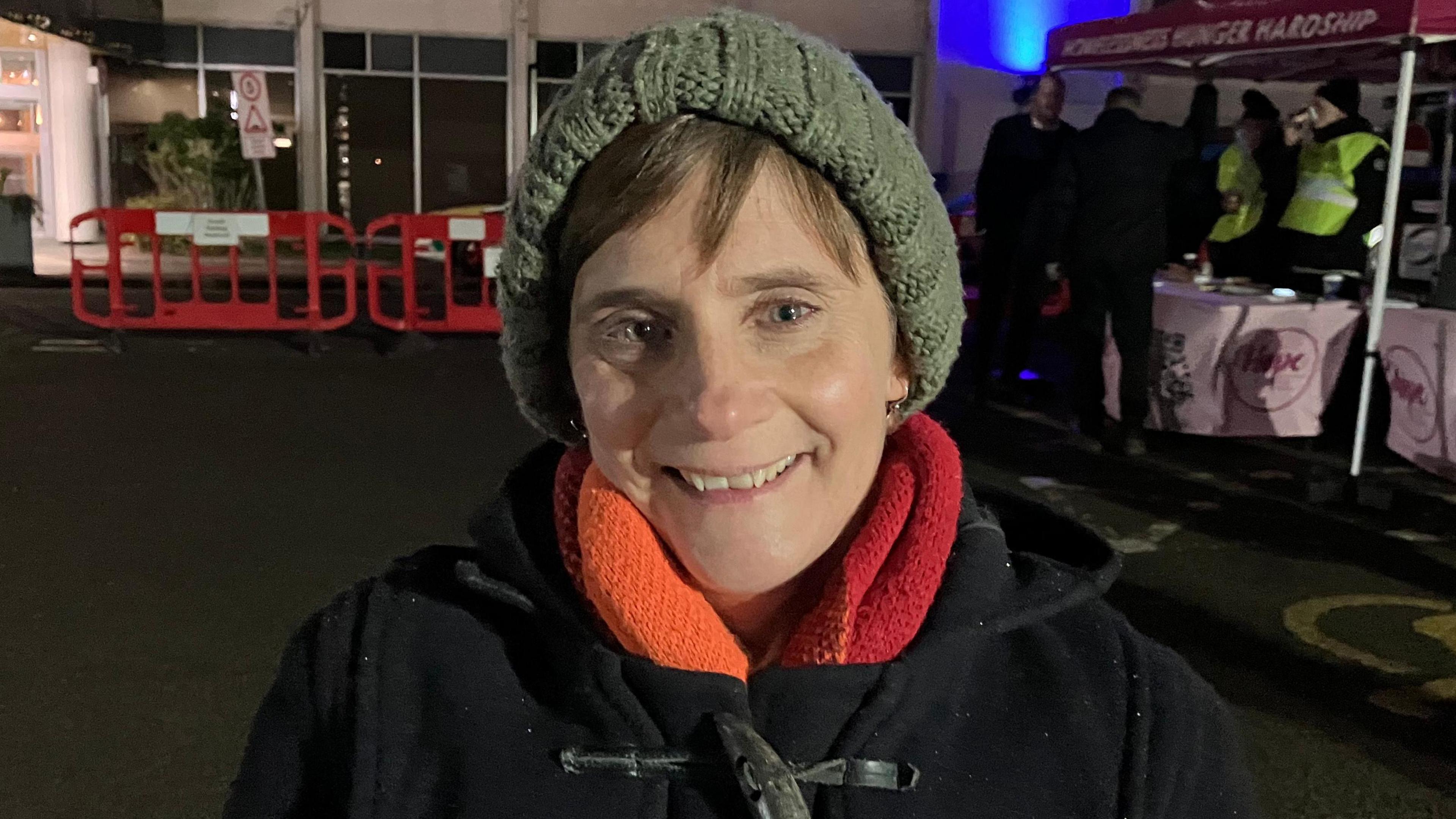 Kyra Williams is seen in the picture wearing a black coat, a red and orange scarf, and a green hat. She is smiling and looking into the camera. 