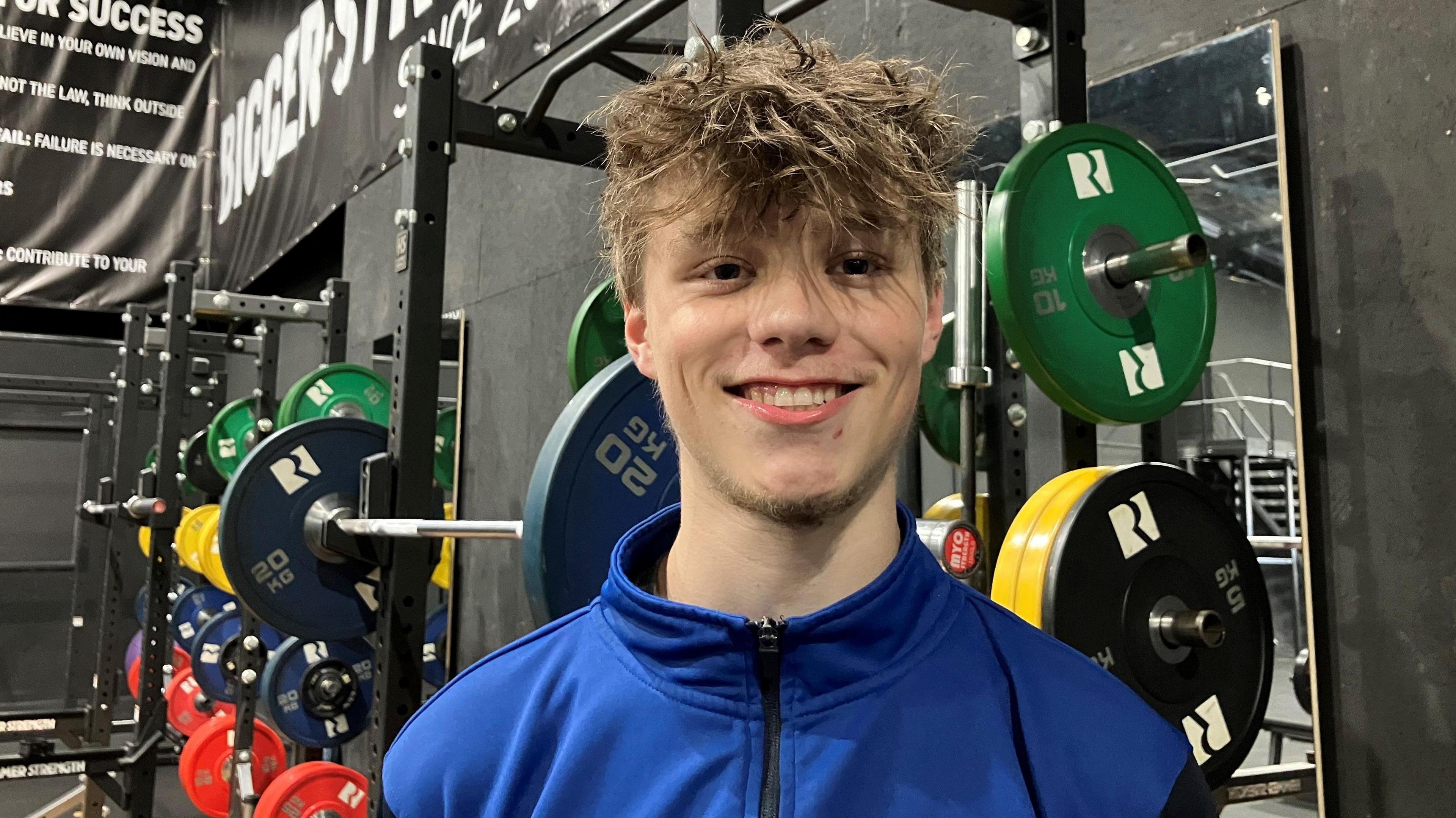 A young man in a blue zip up top standing in a gym in front of a squat rack. He smiles at the camera and has light brown hair. It is a head and shoulders shot.