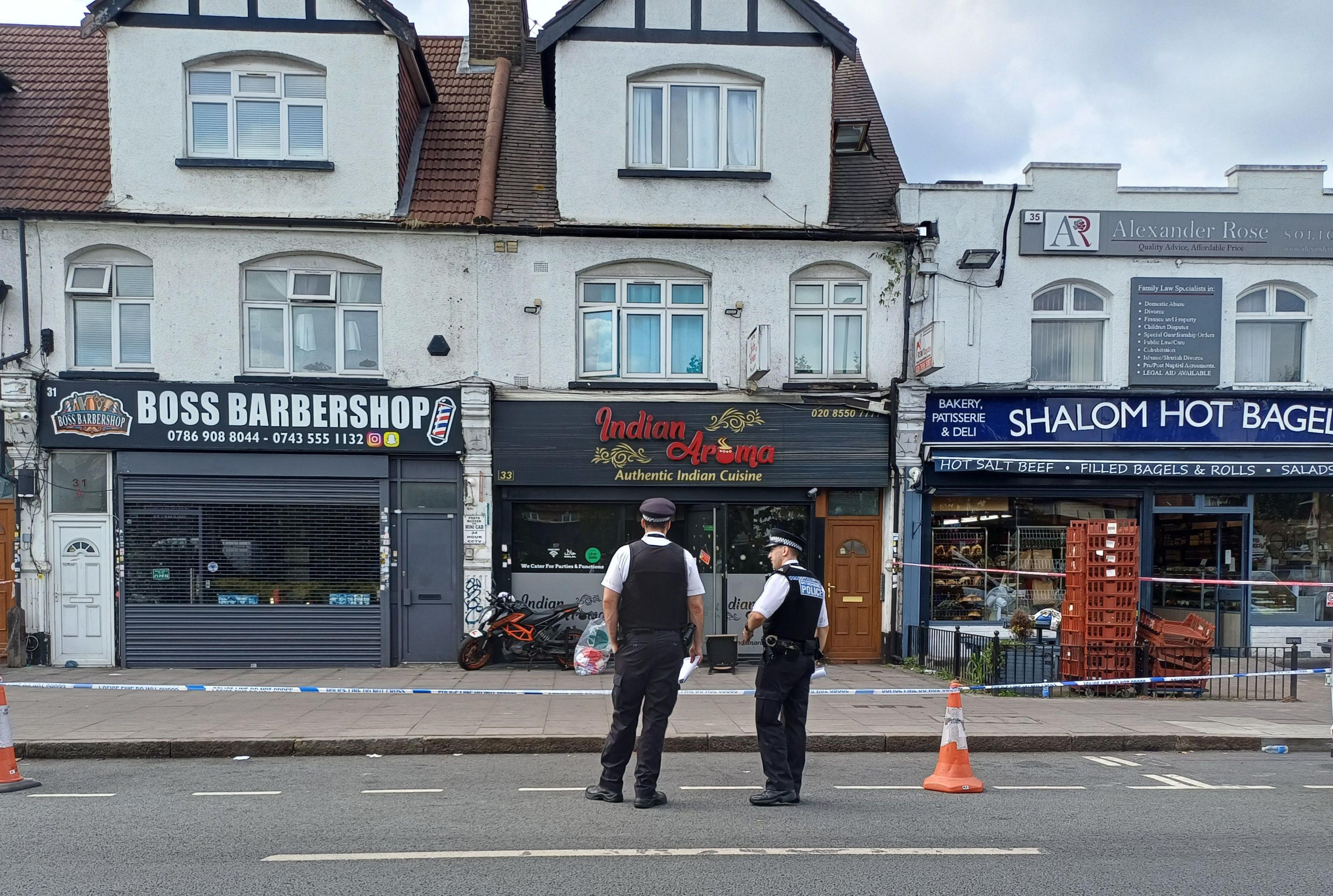 Two police officers stand on a road in front of police tape which surrounds shops and restaurants