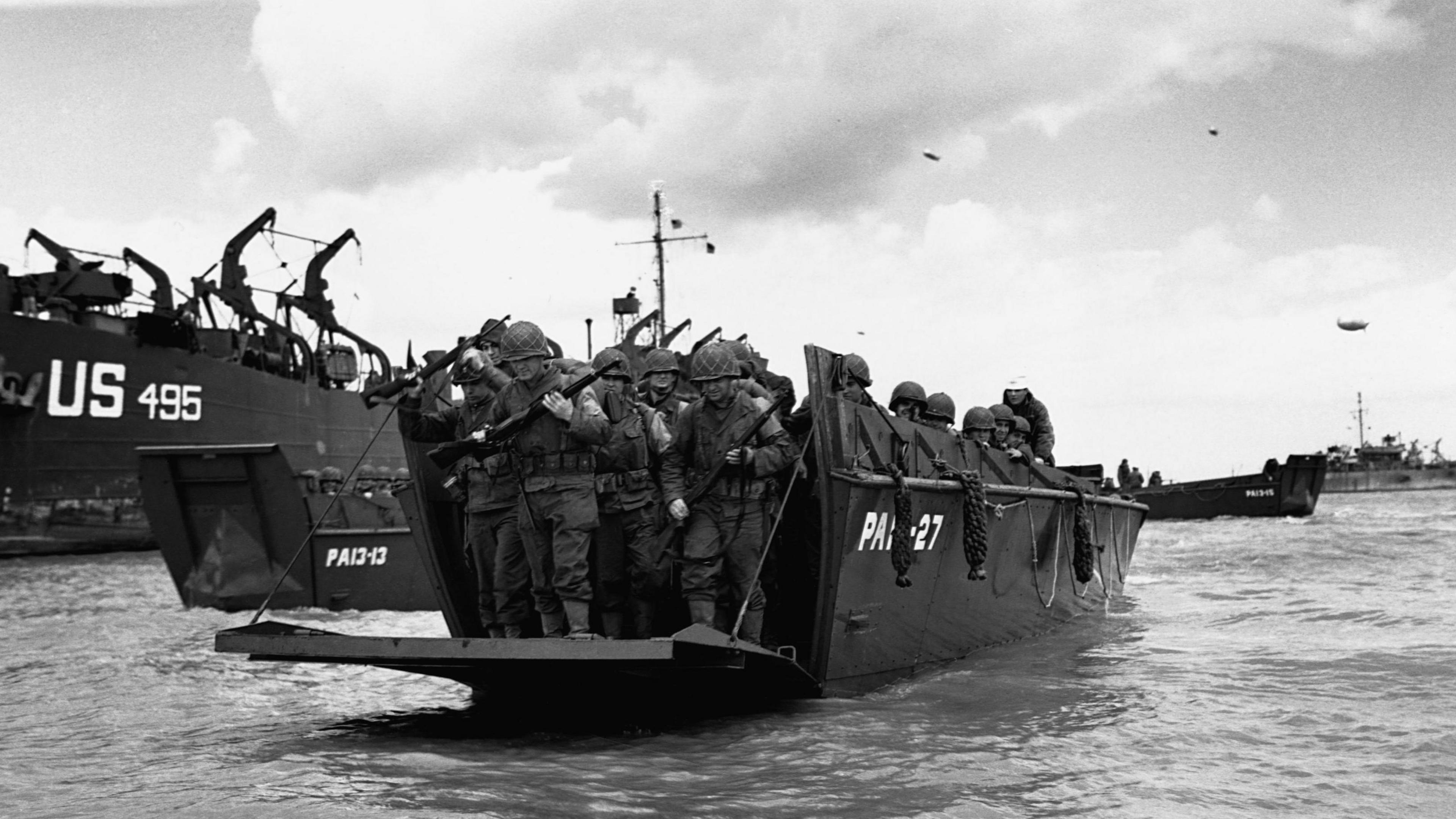 Black and white photo of gun-carrying troops standing on a landing craft. The landing craft is surrounded by sea, and the front of the craft is open. Patches of cloud are seen overhead..