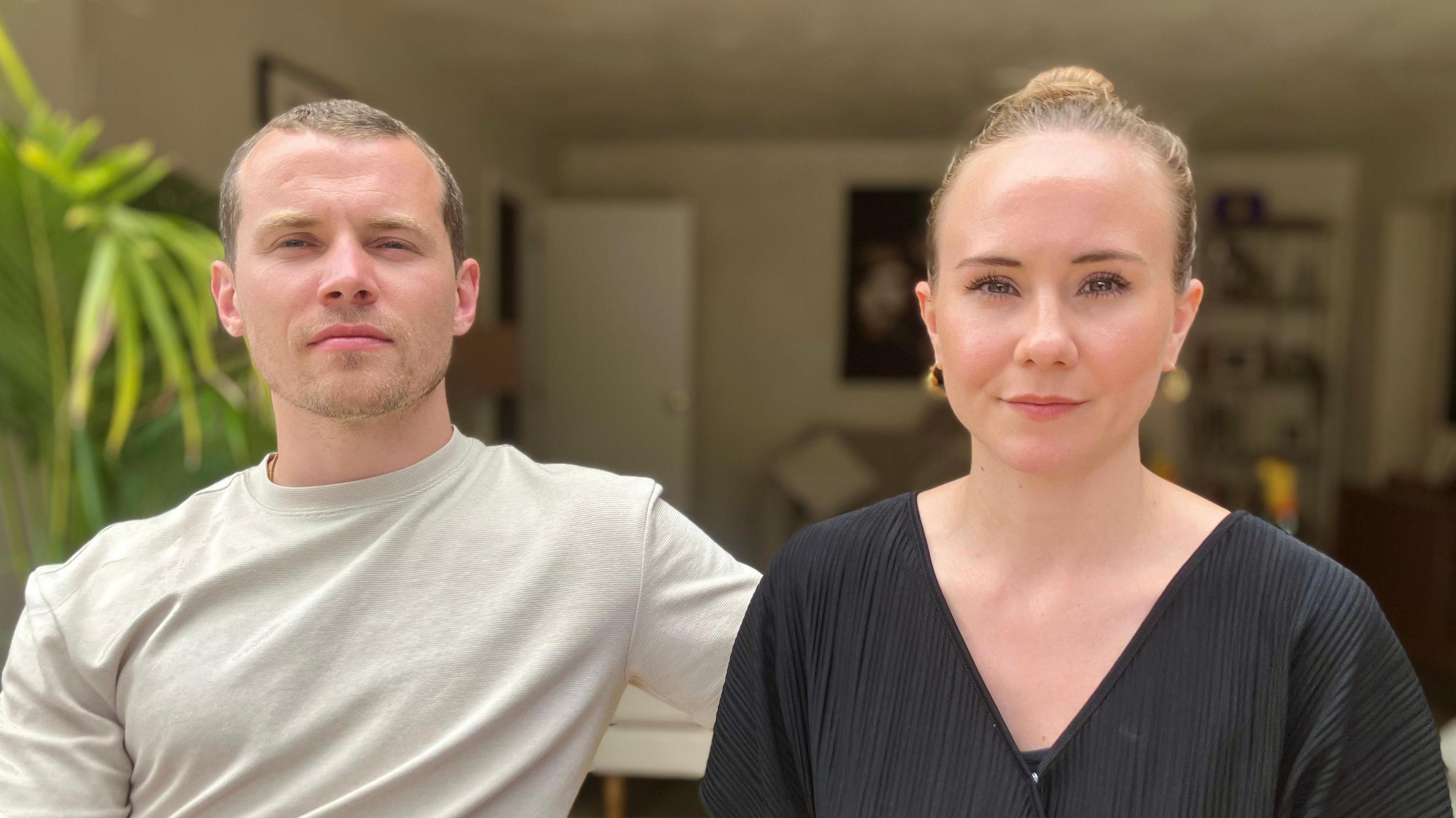 A man and woman sitting together. The man wears a pale T Shirt and the woman a black V neck T Shirt and they are sitting in a living room with plants and pictures on the wall.