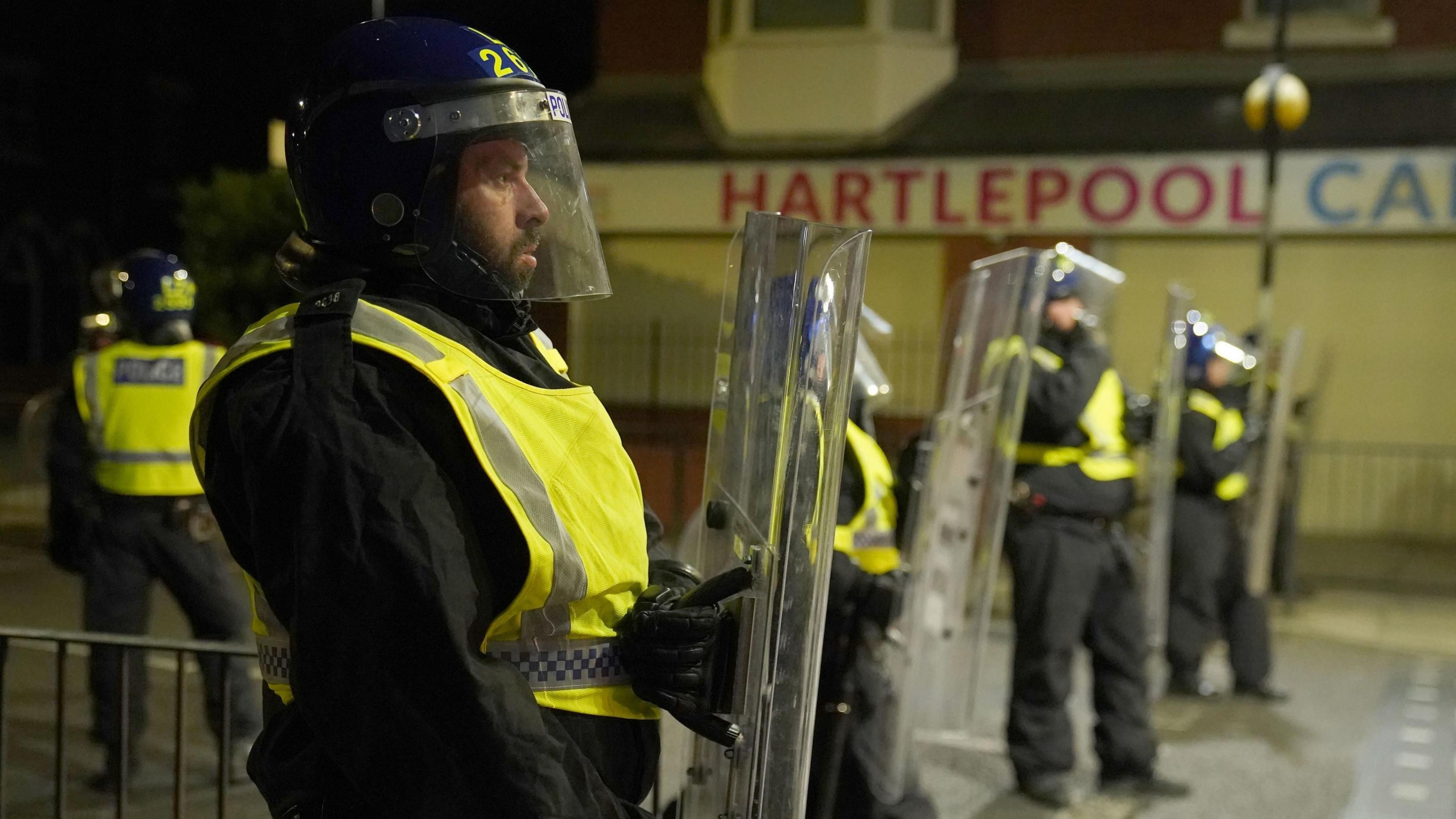 A police officer in a hi-vis vest wears a riot helmet and holds a riot shield. He is standing next to other officers on a road outside a building that has Hartlepool on a sign. It is night-time.