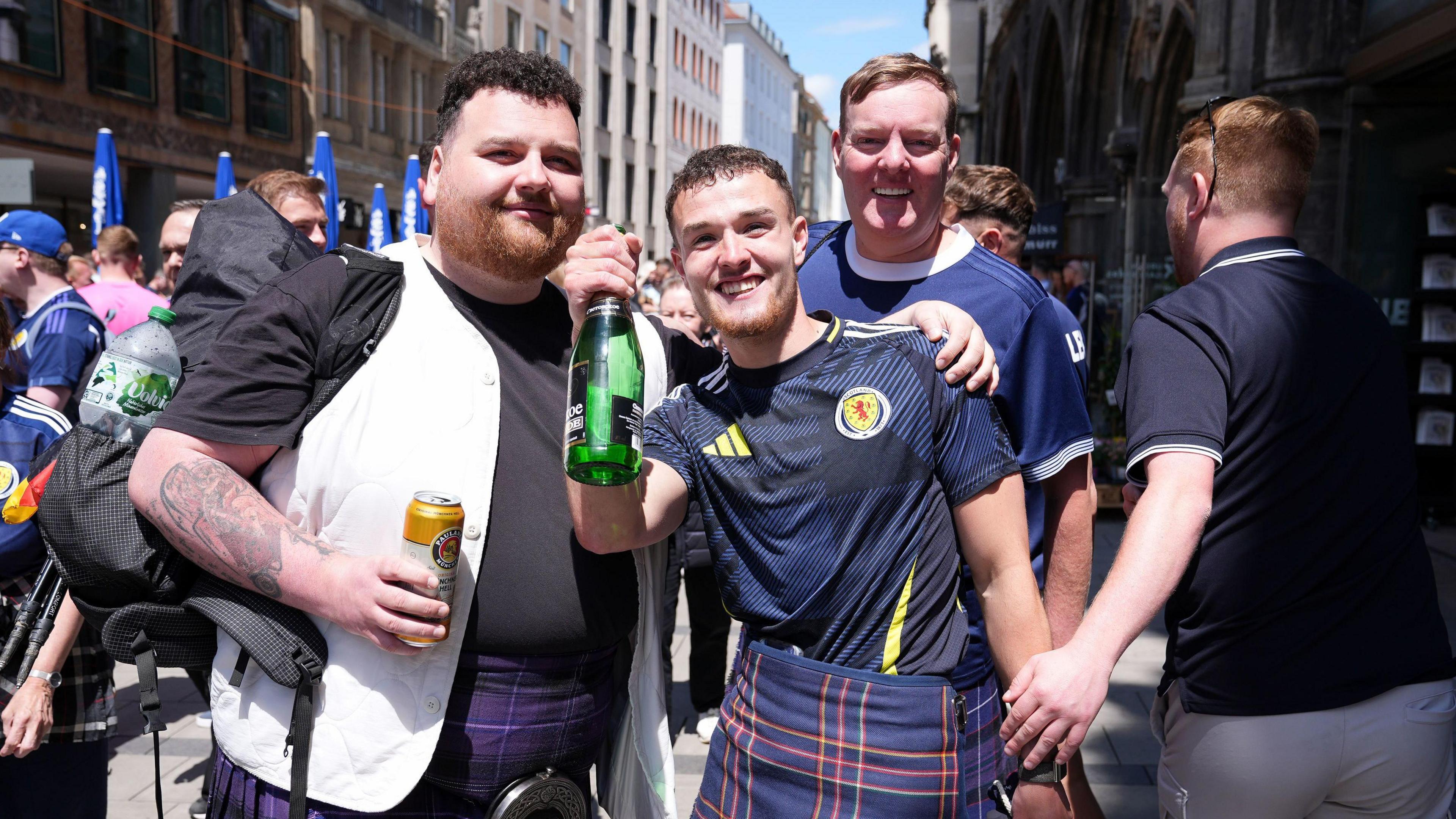 Craig Ferguson in the centre with two friends. He is wearing a Scotland football shirt which is dark blue with yellow and white flashes and a dark blue kilt with red, white and green flashes. He is holding a green bottle.