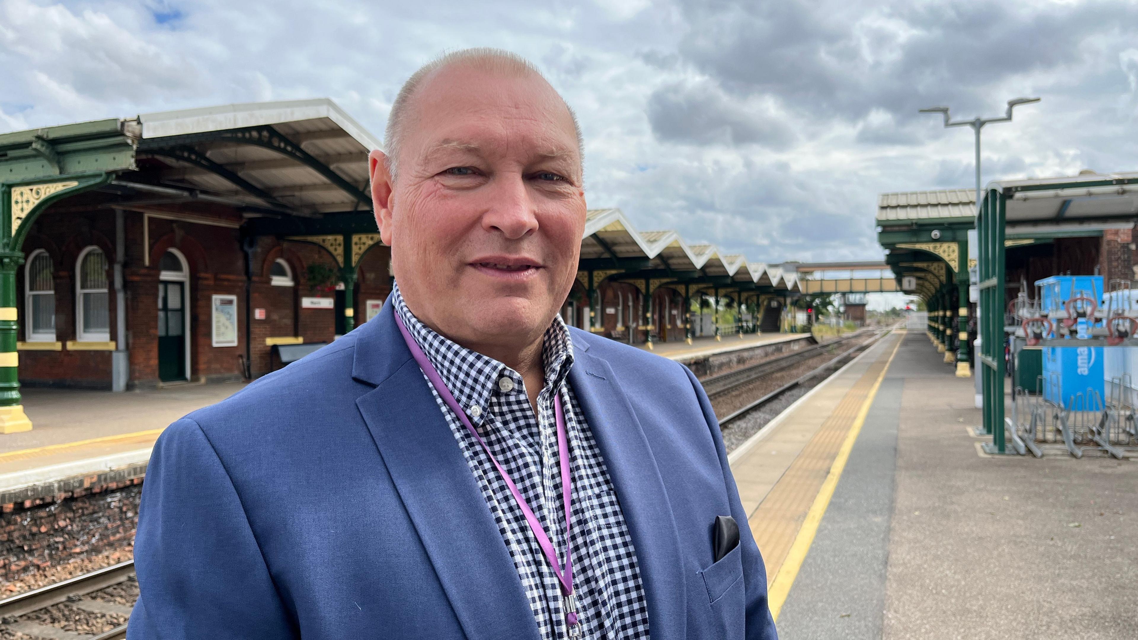 A man standing on a platform at a railway station. He has very short fair hair and is wearing a blue and white check shirt and a blue jacket. Behind him are railway tracks and two platforms.