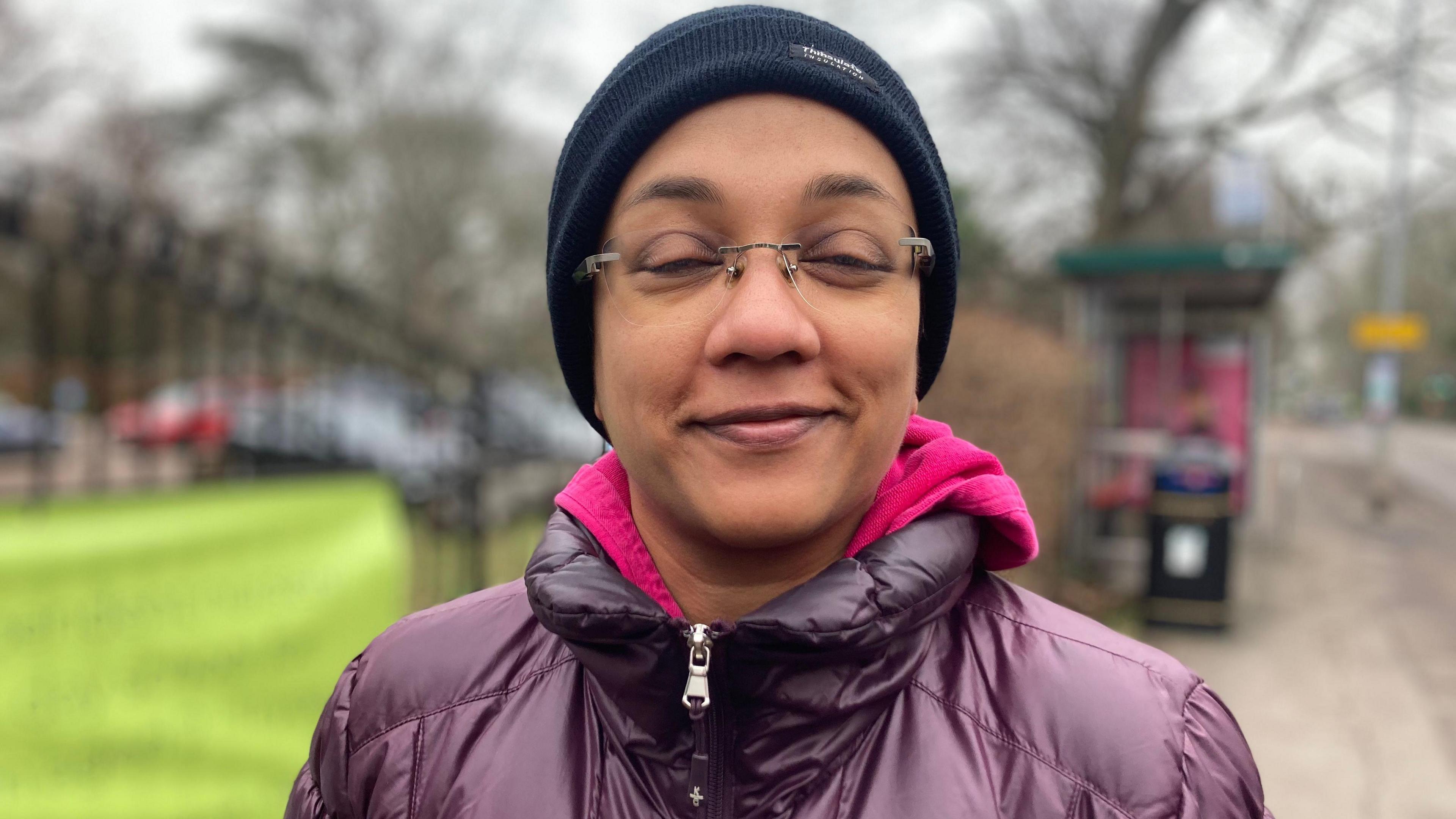 Pranathi Ramachandra wearing a winter woollen hat and a warm purple jacket and pink jumper. She smiles and there are some park gates behind her. 