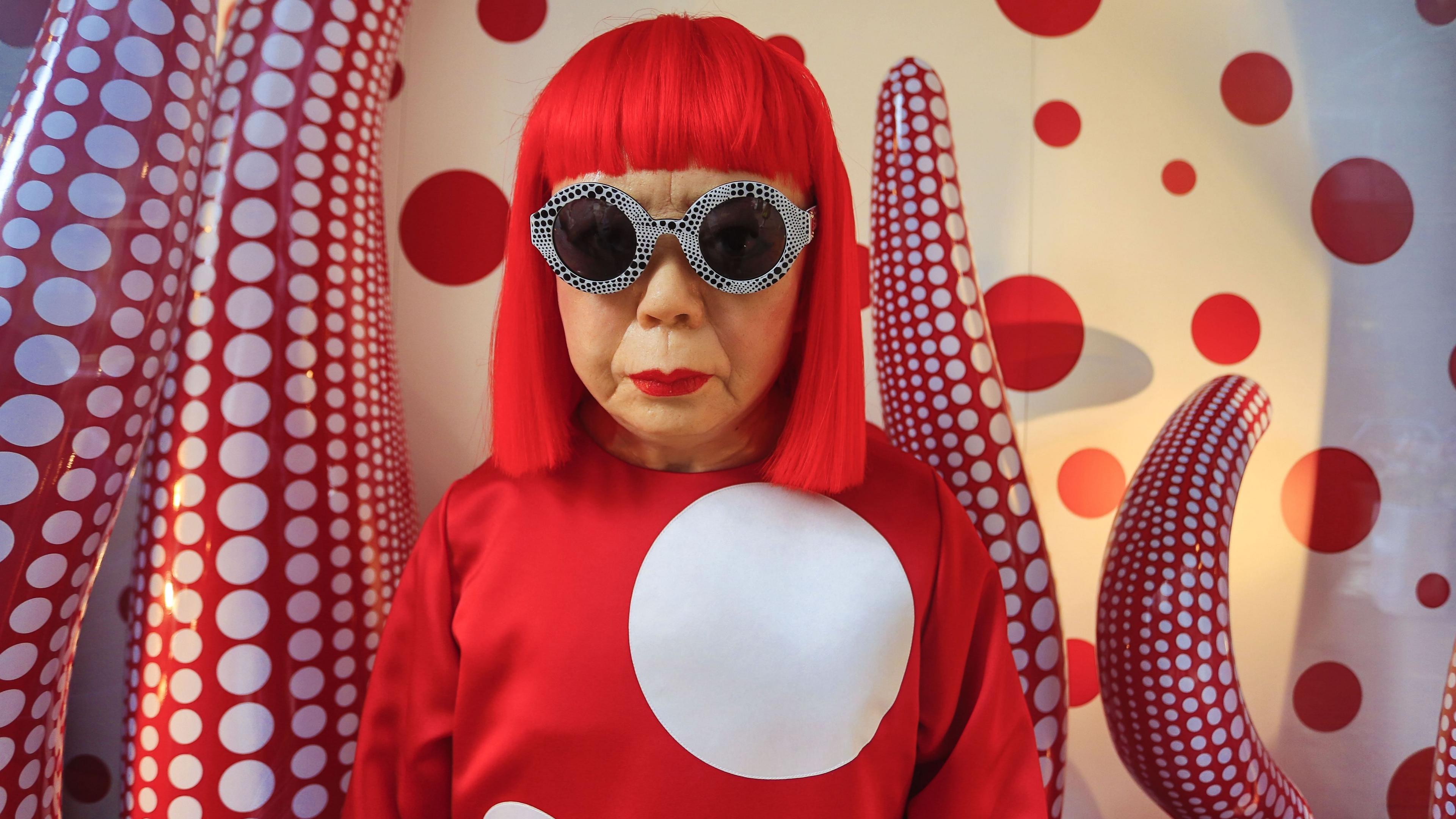 Yayoi Kusama with red wig, red top with a large giant spot and goggles, in front of a red and white spotty tentacle-like artwork.