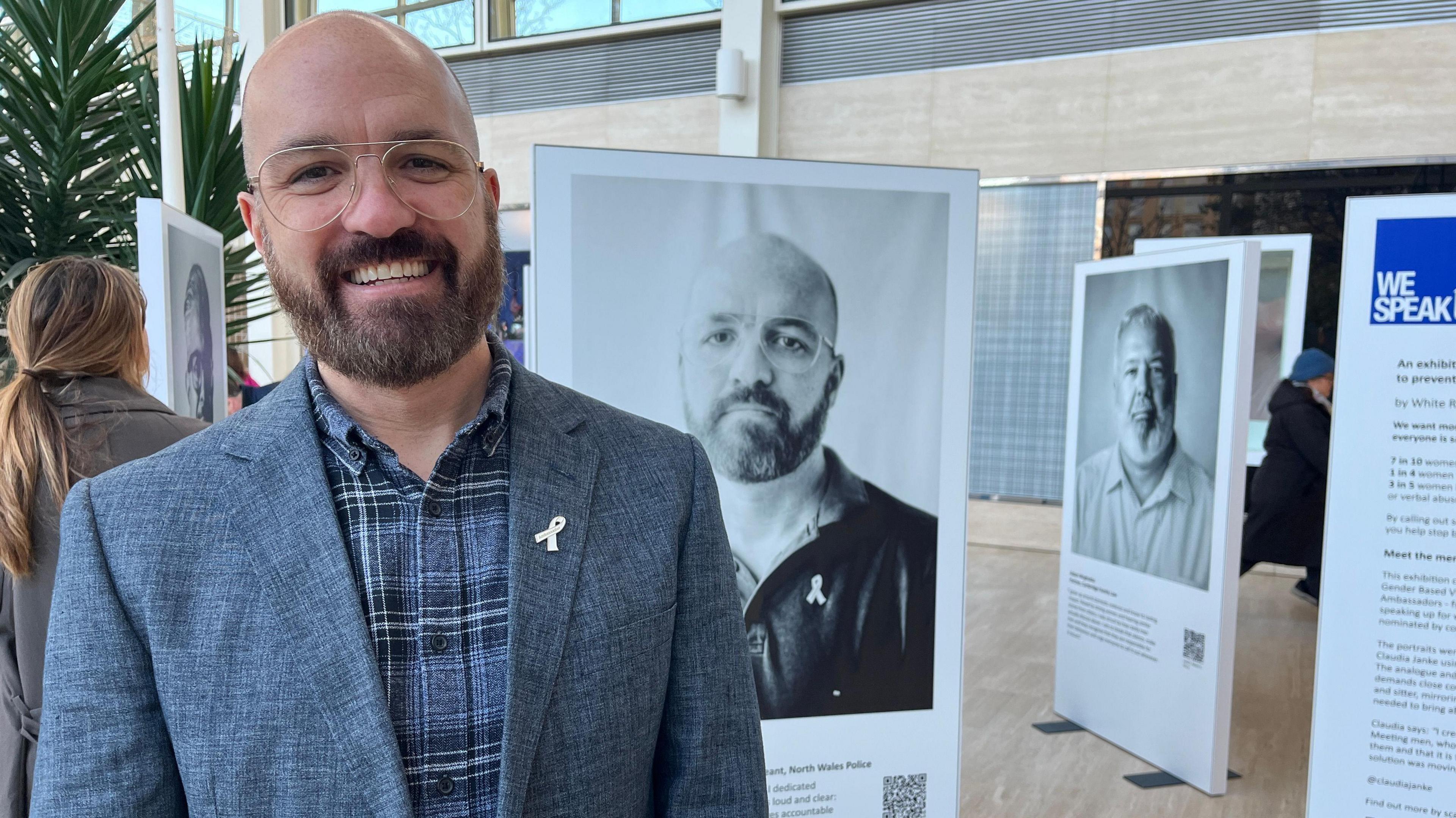 Mike Taggart also stands in front of the portrait of him that features in the exhibition. He is bald and wears glasses. He is wearing a blue-checked shirt underneath a grey jacket, which has a white ribbon pinned to the lapel. He is smiling. Other portraits in the exhibition can be seen behind him.
