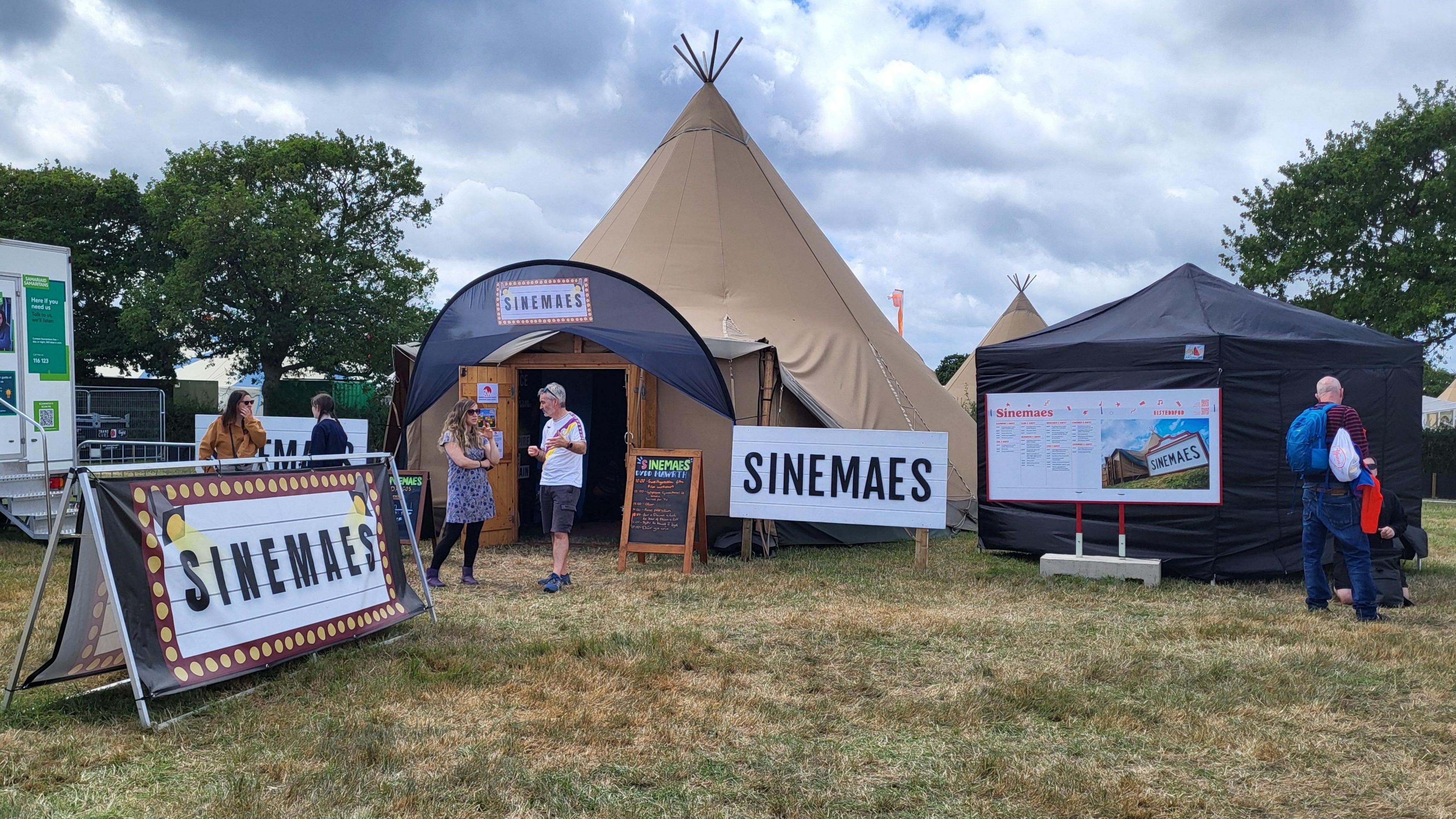 Brown cinema tent with a huge sign saying Sinemaes. There are a couple of people stood in front of the tent. 