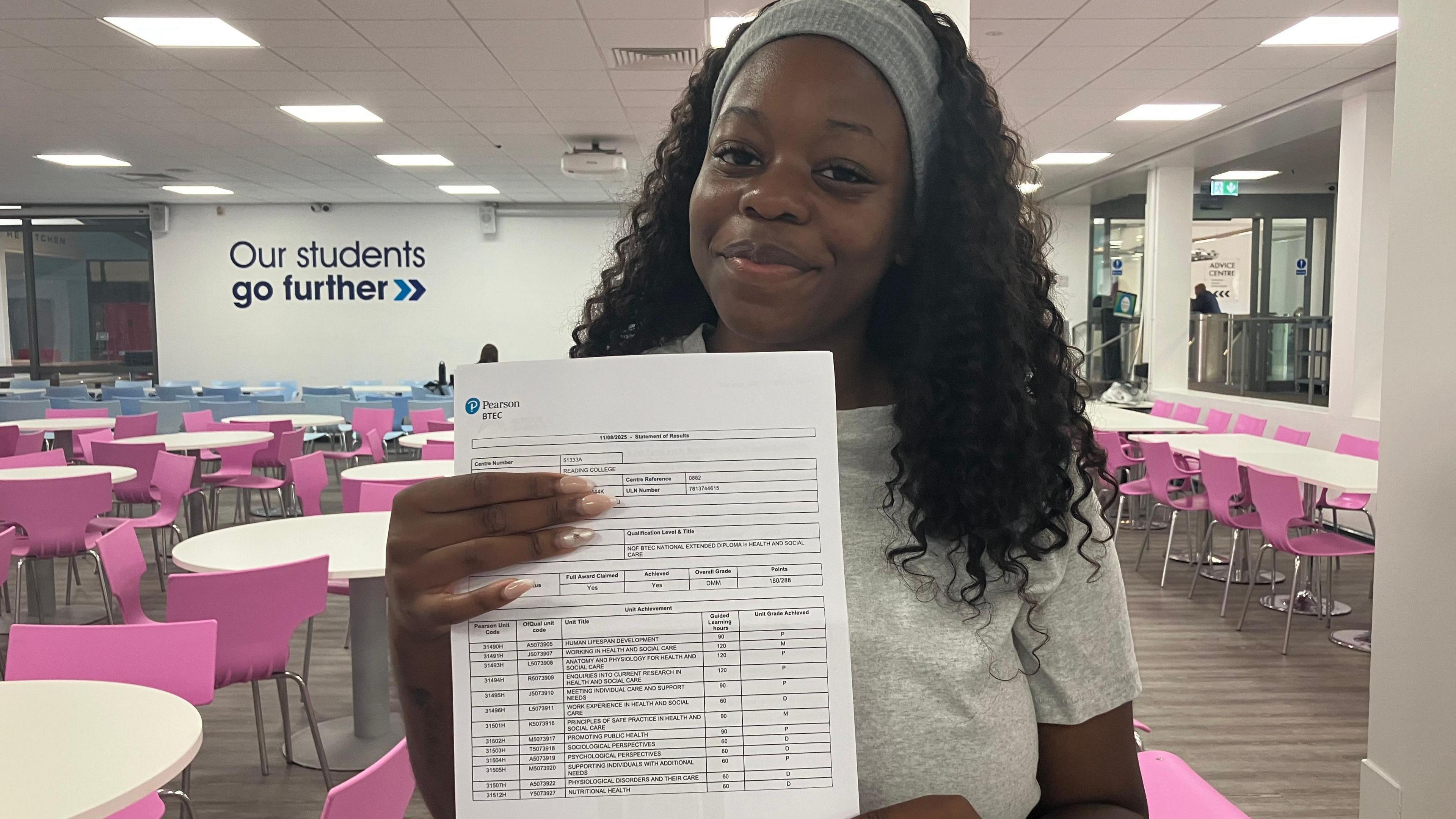 Tawana Mandebvu stands in her college canteen, holding the paper with her results up to the camera. She is smiling. Tawana has long black curly hair and wears a grey elastic headband to keep her hair back. She has a light grey short sleeved t shirt on. Behind her are a number of white tables with pink chairs. 