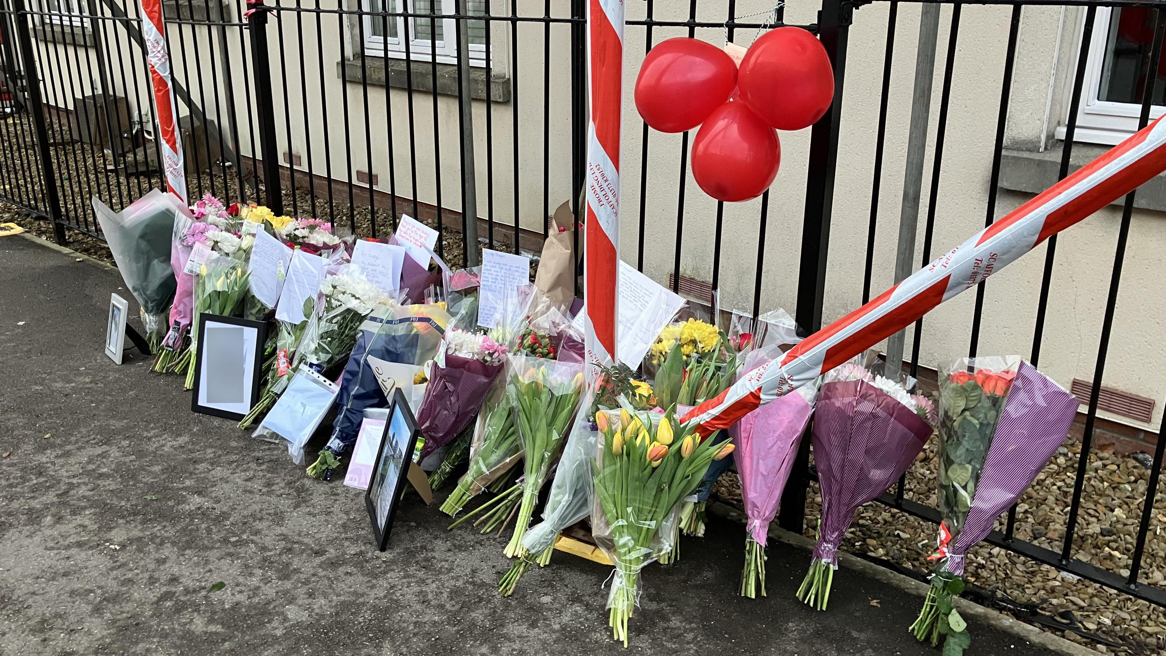 Dozens of bunches of flowers placed against a set of railings on London Road in Chippenham. The flowers include tulips, roses and carnations and some have messages pinned to them. Above the bunches of flowers are three red balloons tied to the railings. 