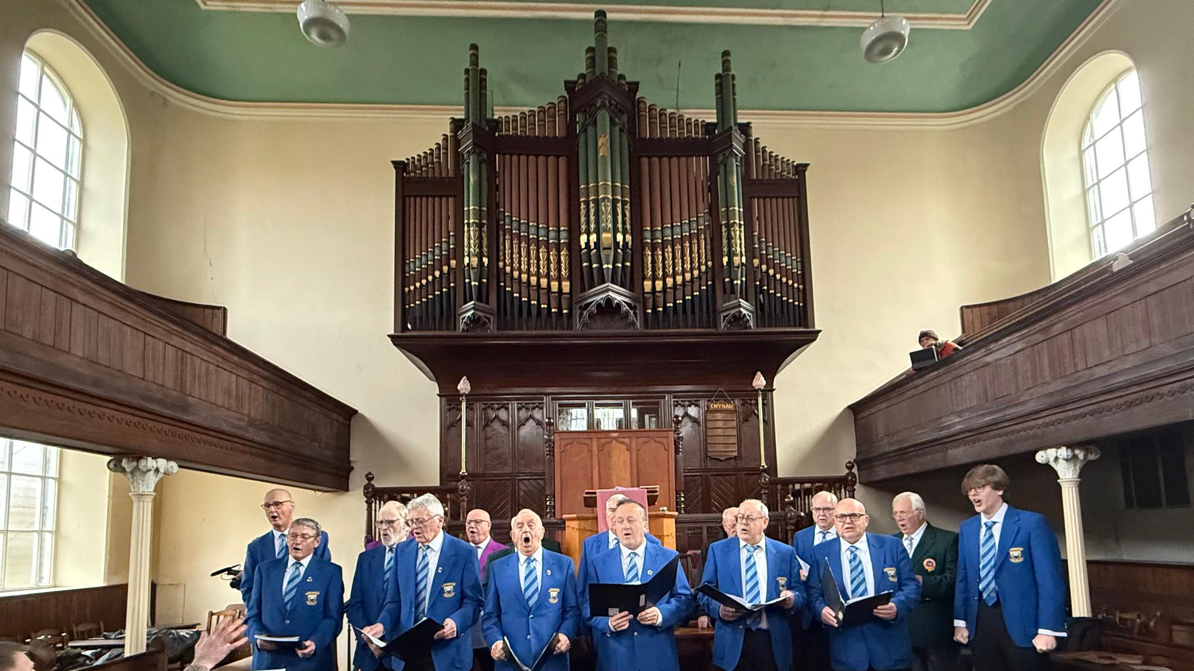 A male voice choir dressed in blue blazers and blue ties and holding choral hymn books sing in front of a large ornate pipe organ. 