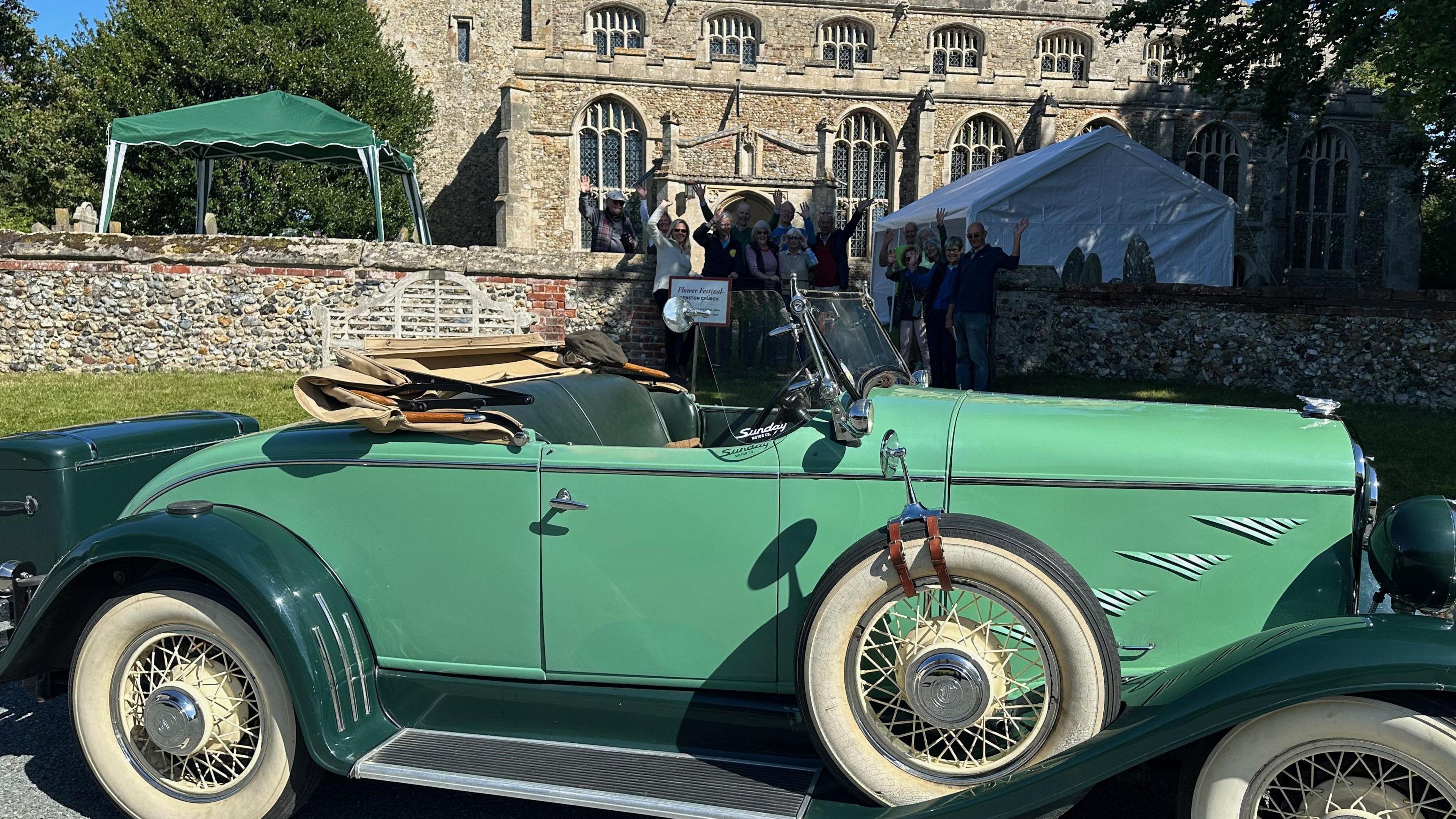 A green coloured vintage car is pictured parked outside a church. In the background a group of people are gathered and smiling at the camera. A few have their arms raised in the air in celebration. A church can be seen behind them.