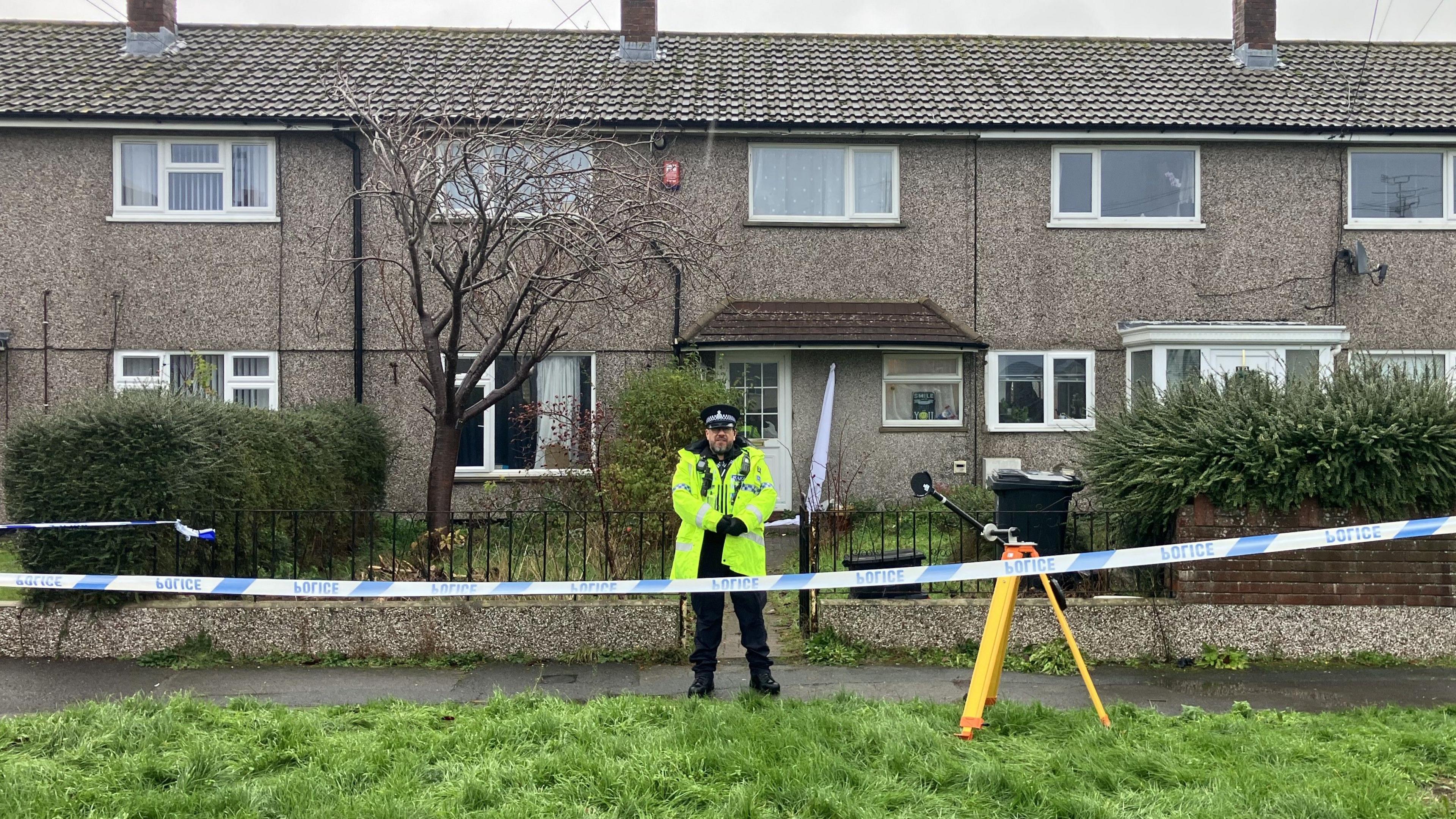 A policeman dressed in a yellow hi-vis jacket, black trousers and police cap standing outside the home where the incident occurred. It is a grey pebbledash two-storey home with a front garden and black railing gate. There is blue and white police cordon tape stretching across the front of the property.