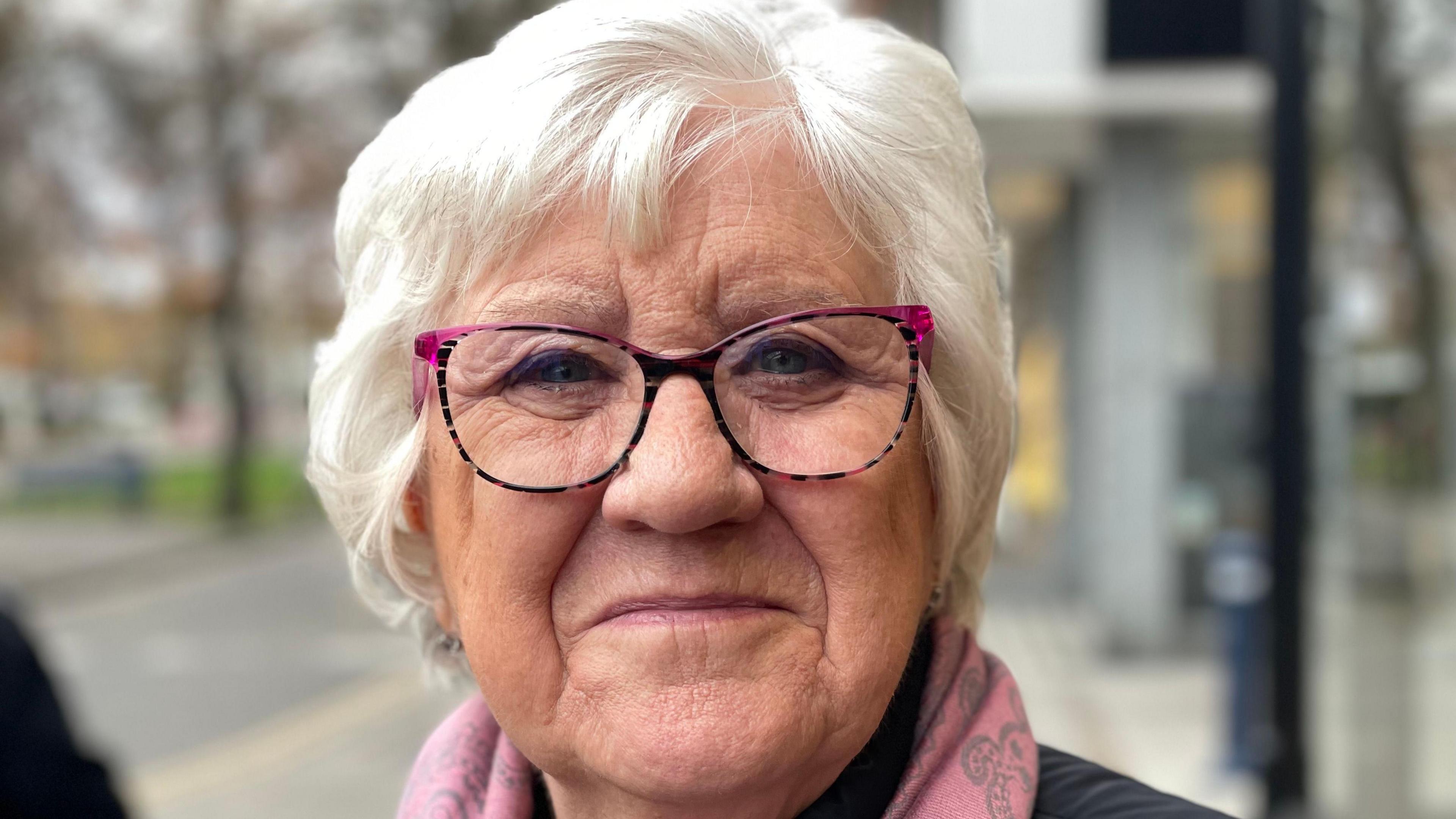 Hilda who has grey hair and wears horn rimmed glasses and a pink scarf smiles at the camera.