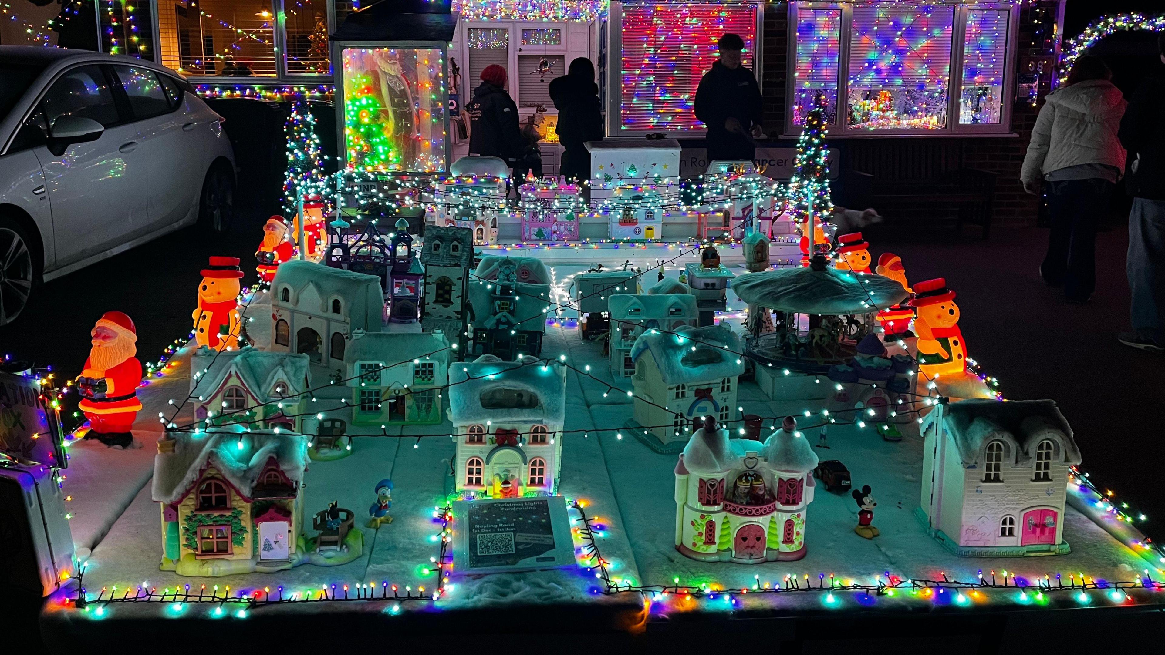 A model village is surrounded by its own mini fairy lights and includes snowmen, Father Christmas figurines and a toy train. Multicoloured lights are visible in the background. 