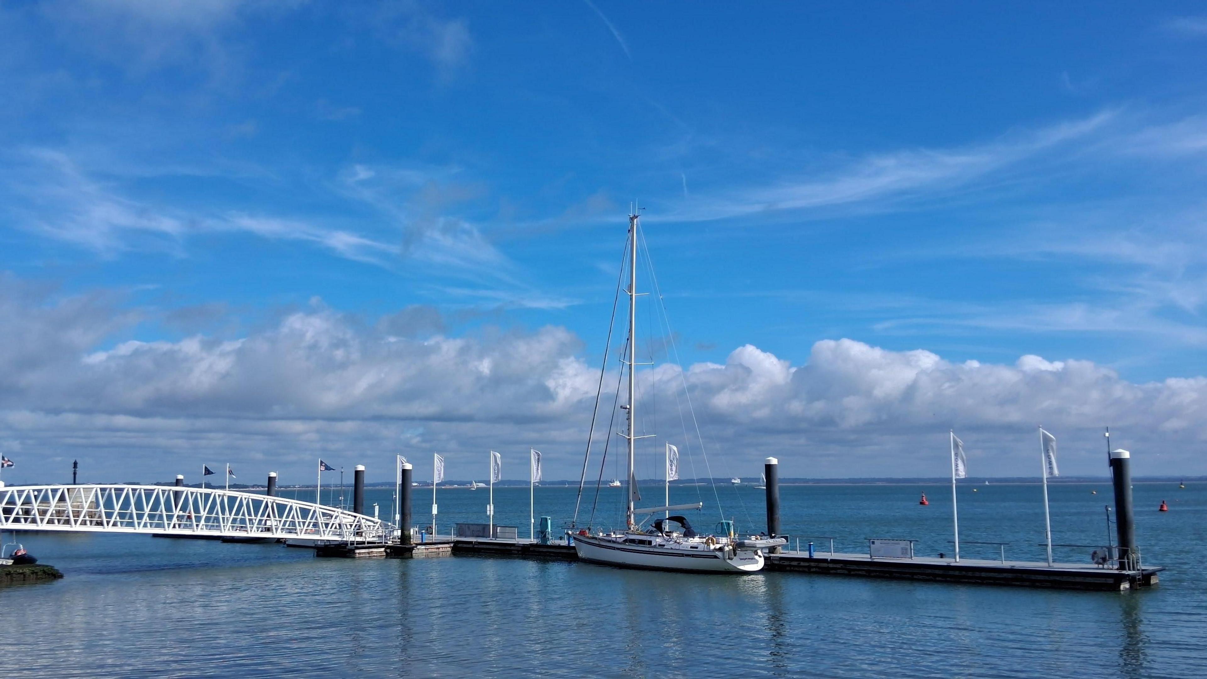 A white yacht is moored next to a pontoon and a white walk-way bridge. The sea is blue on a sunny day with blue skies overhead and fluffy white clouds.