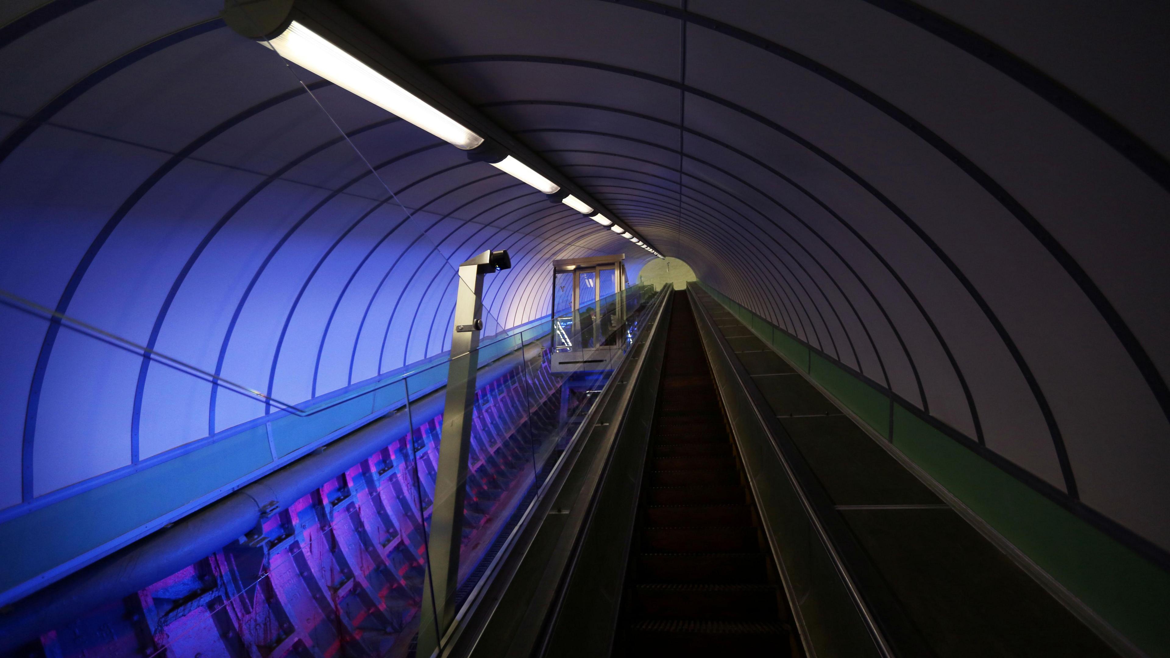 A glass lift travelling along an incline. It has a steel framework. The tunnel area is illuminated.