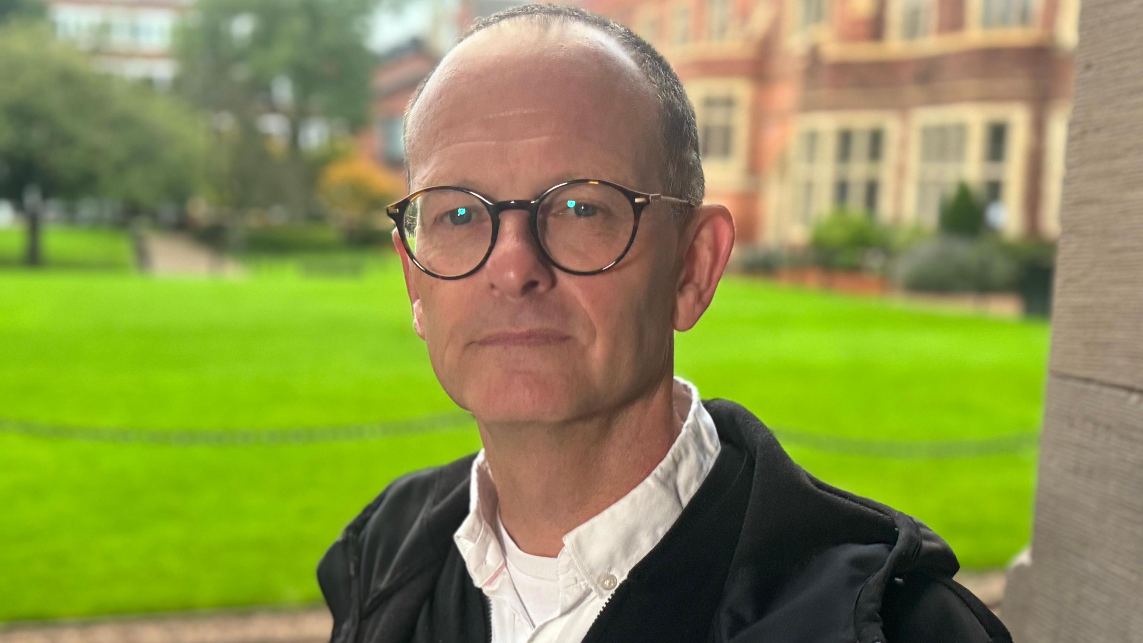 Professor Geraint Ellis looks at the camera. He is wearing a black jacket, white shirt and round glasses. 