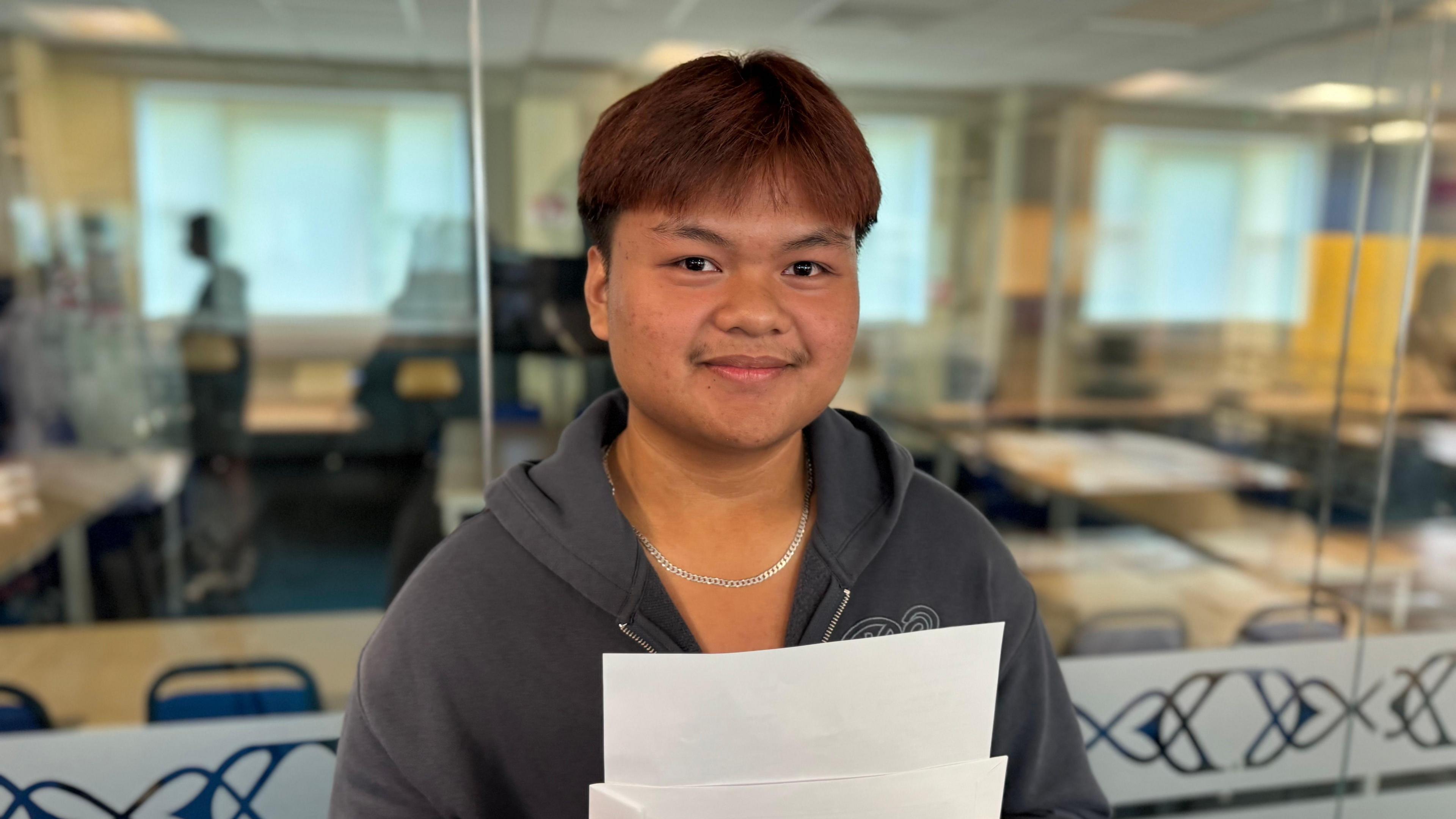 Jhay Lordviloria stands in front of glass classroom windows. He holds up a white piece of paper and envelope. He has short dark hair which has been coloured red and wears a navy blue hooded jumper and silver chain around his neck.