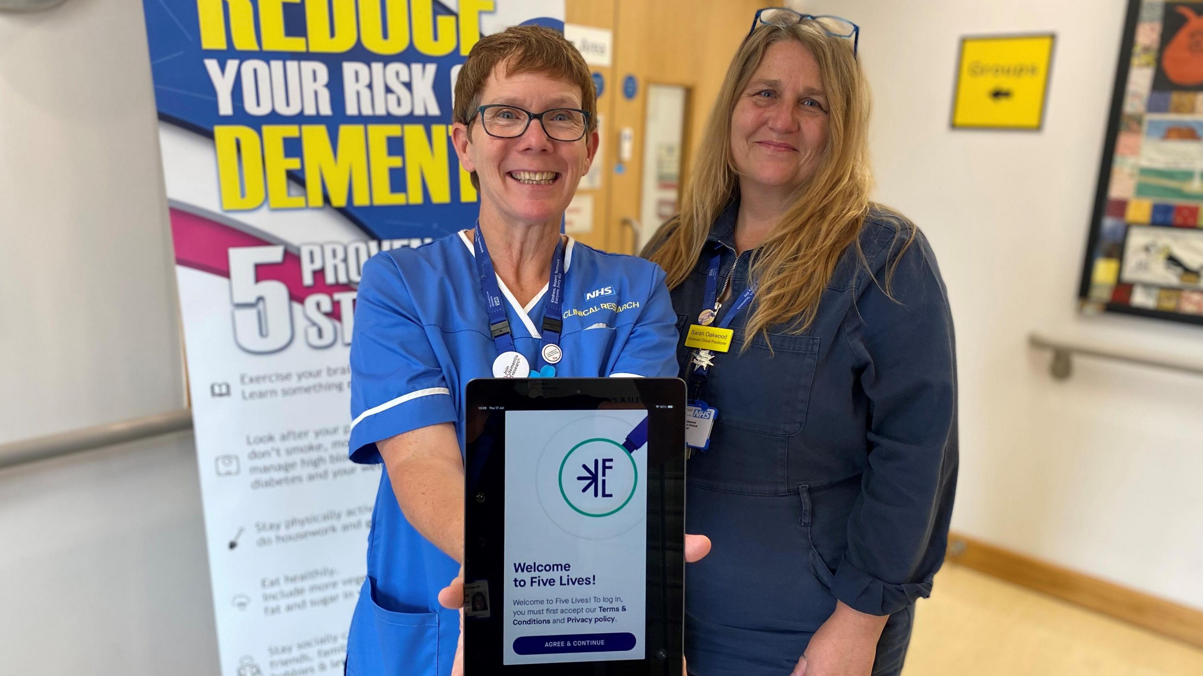 Two women in blue scrubs are standing in a hospital hallway smiling at the camera. They are holding up a tablet which says "Welcome to Five Lives".