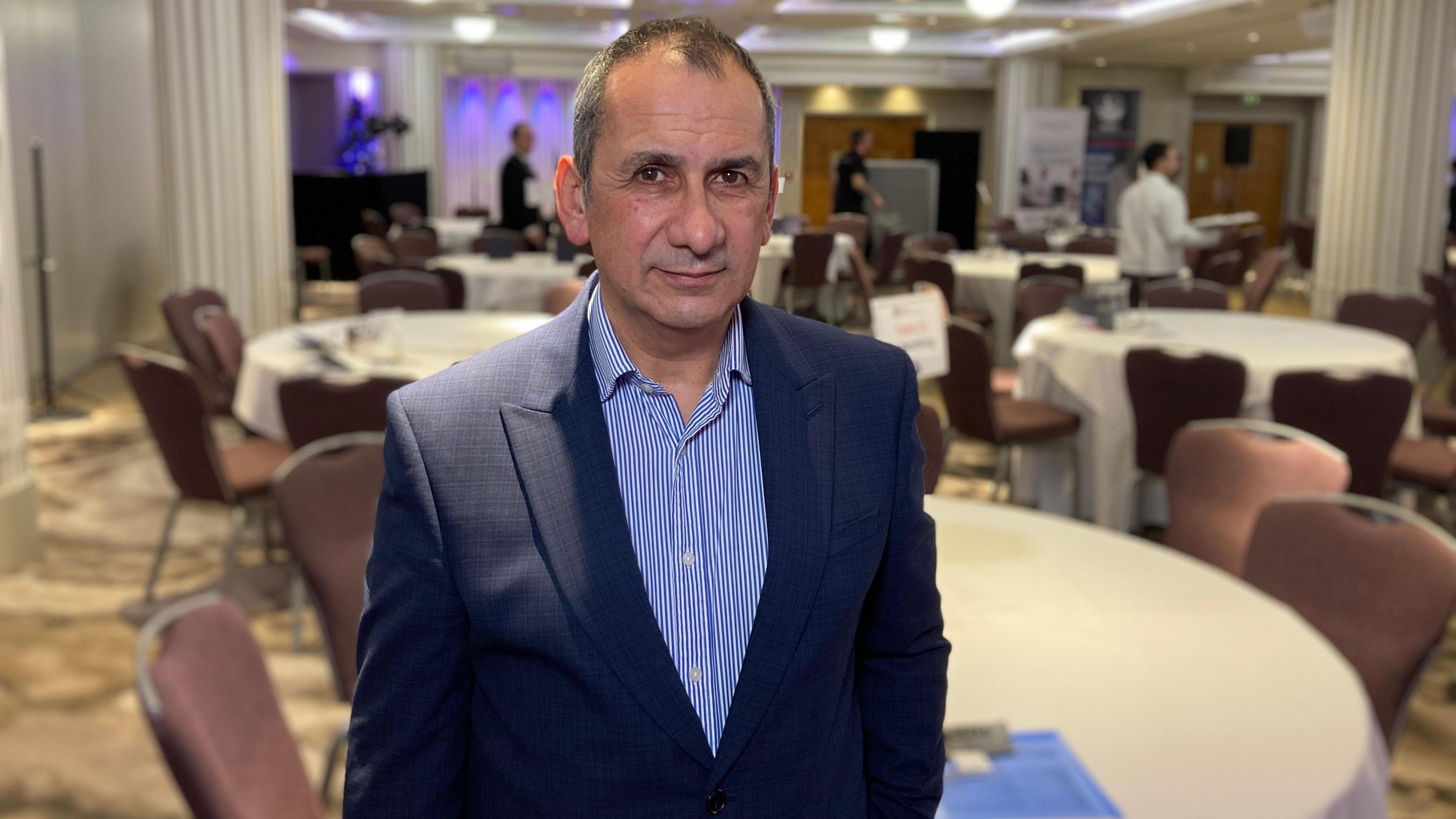 Murray Norton wears a blue striped shirt and blazer. He is standing in a room with round tables and beige chairs.