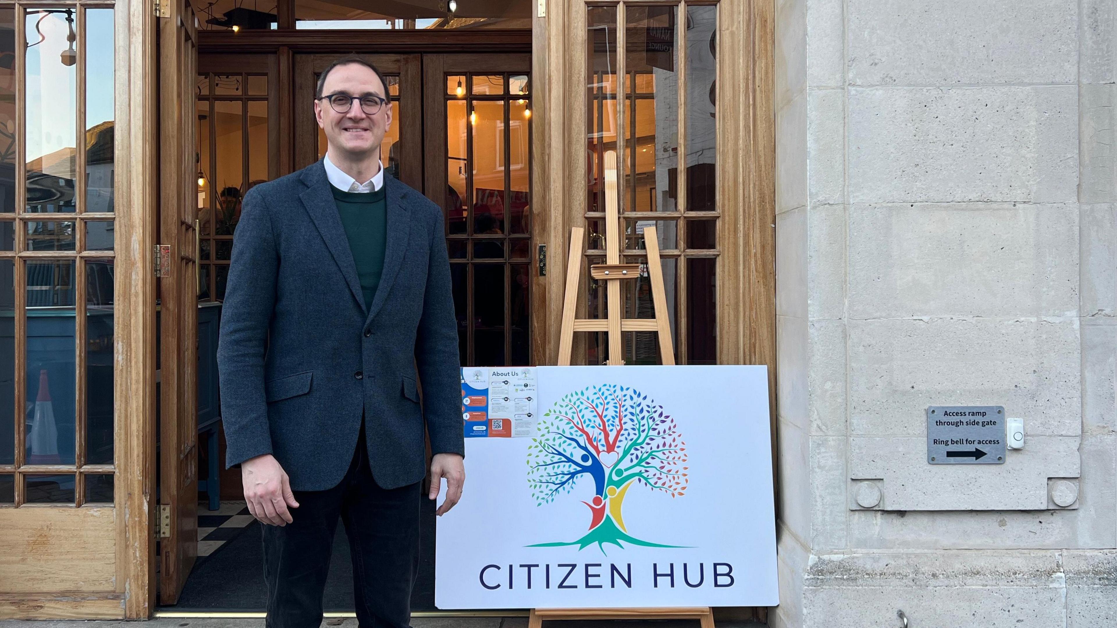 Ian Sollom standing outside the new hub next to a white sign which reads 'Citizen Hub'. He is wearing a suit, circle-framed glasses and a shirt