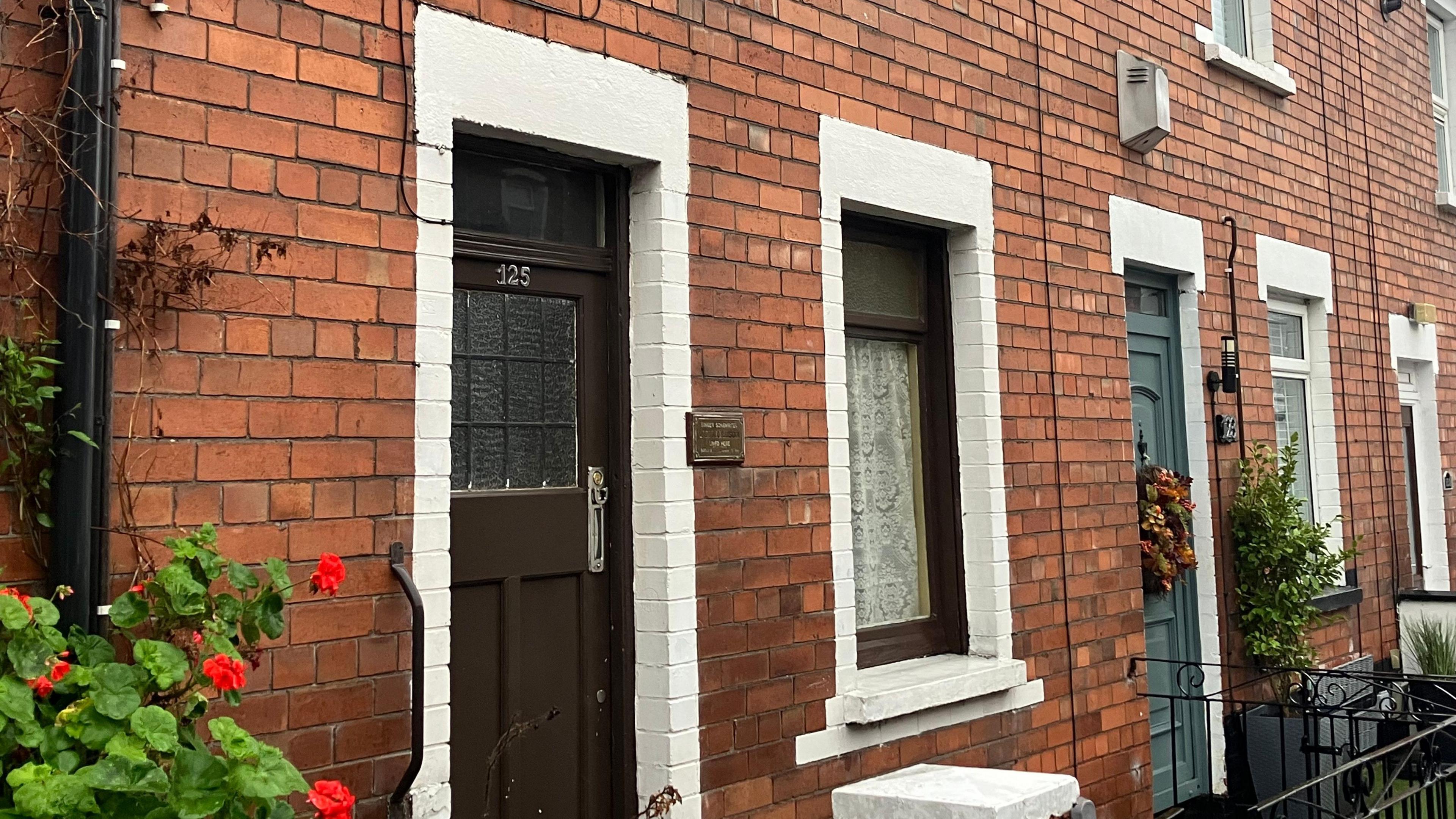 A terraced red bricked house. Some red flowers are planted. The door and the window frame are painted brown. 125 is displayed on the door.