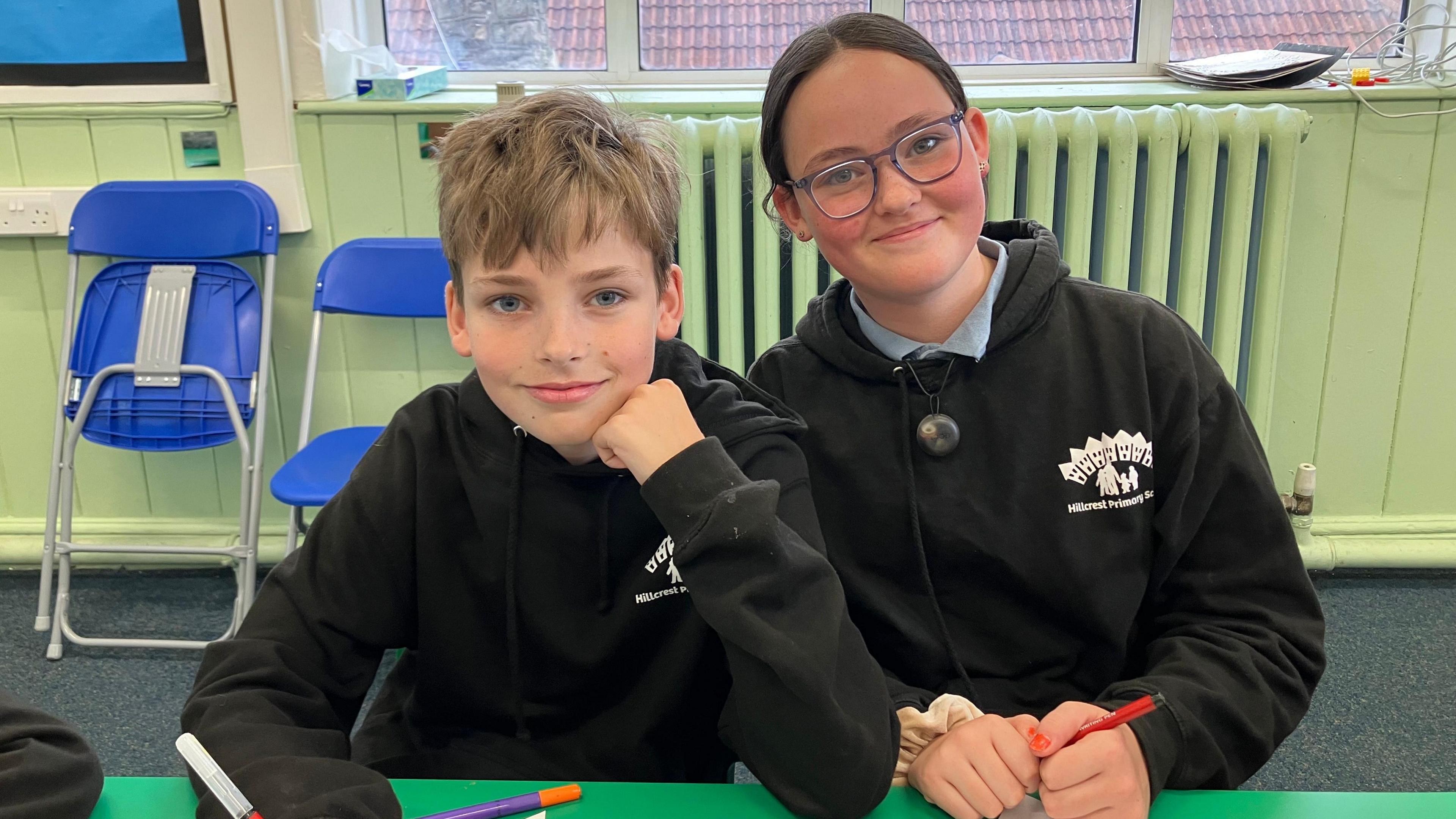 Twin Issac, who has short light blonde hair, sits next to Eva, who has brown hair tied back in a pony tail and purple glasses. Both are wearing school black leavers hoodies, smiling