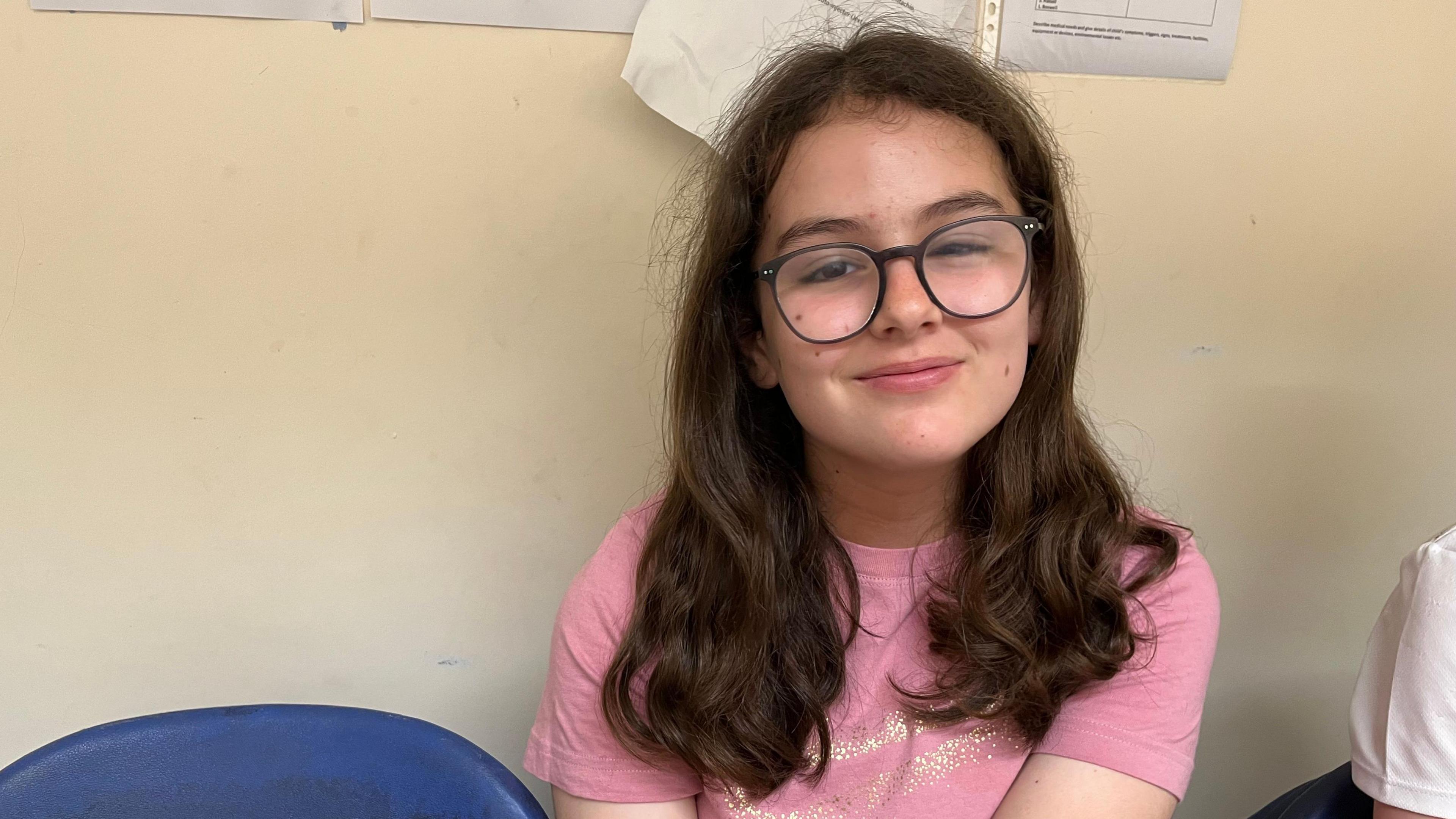 Freya has long dark-brown hair and glasses and is wearing a pink t-shirt. She is sitting on chair with a light-coloured wall behind her.