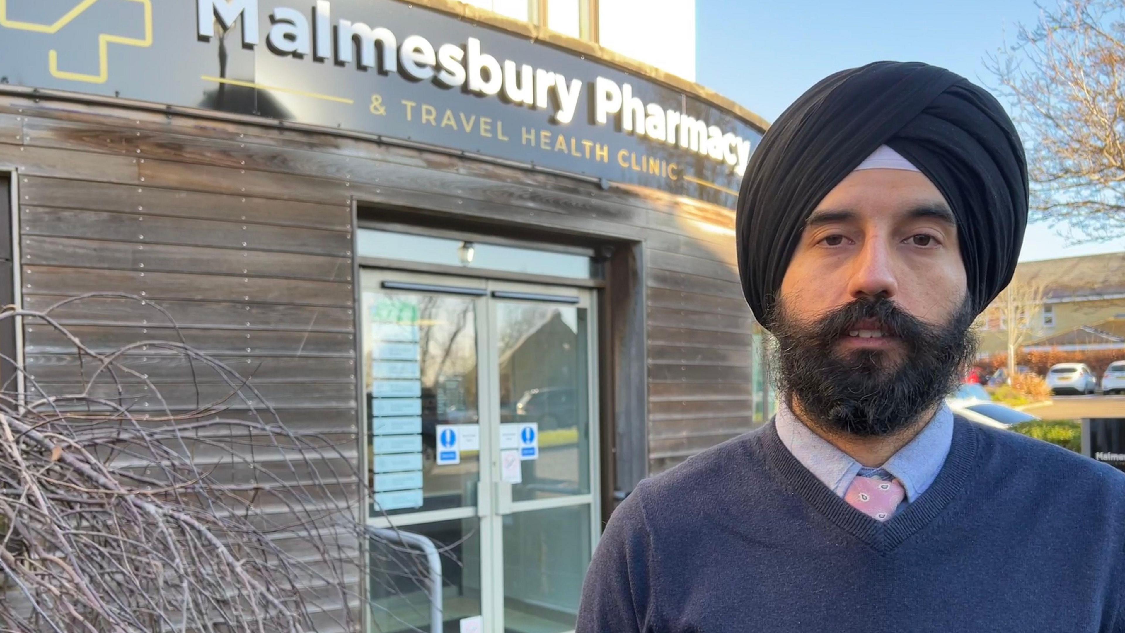 Gurinder Singh pictured outside his pharmacy. He has a large dark beard and moustache, and is wearing a dark blue jumper, blue shirt, pink tie and a black turban. The pharmacy has a large black and yellow sign reading 'Malmesbury Pharmacy and Travel Clinic". 