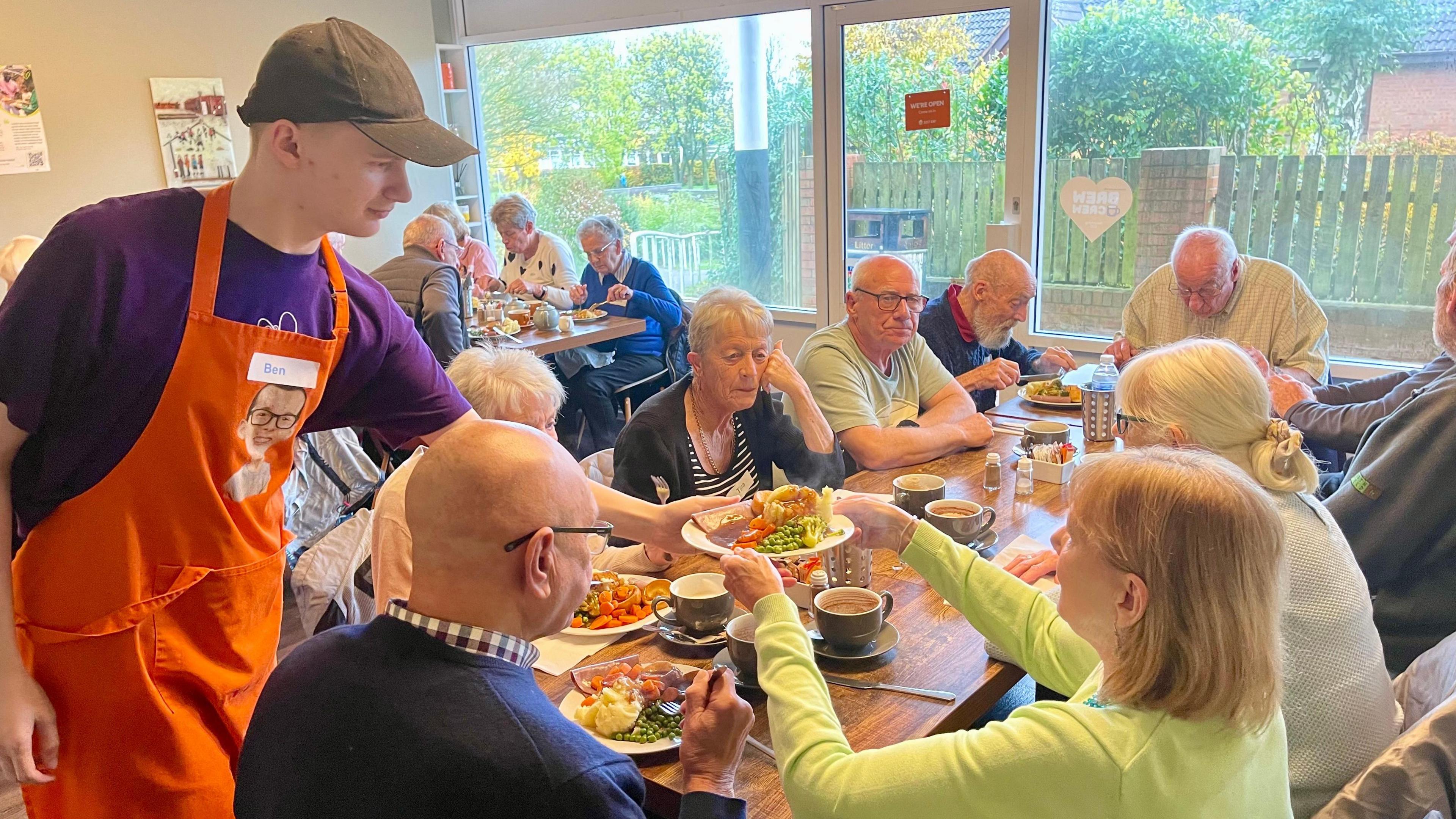 A man in a purple t-shirt, orange apron and black baseball cap is handing a full roast dinner to a woman in a lime green cardigan. She is sat at a cafe table with nine other men and women about to eat the same meal. There is another table of four people in the background eating roast dinners. The cafe has large windows and a glas door at the front.