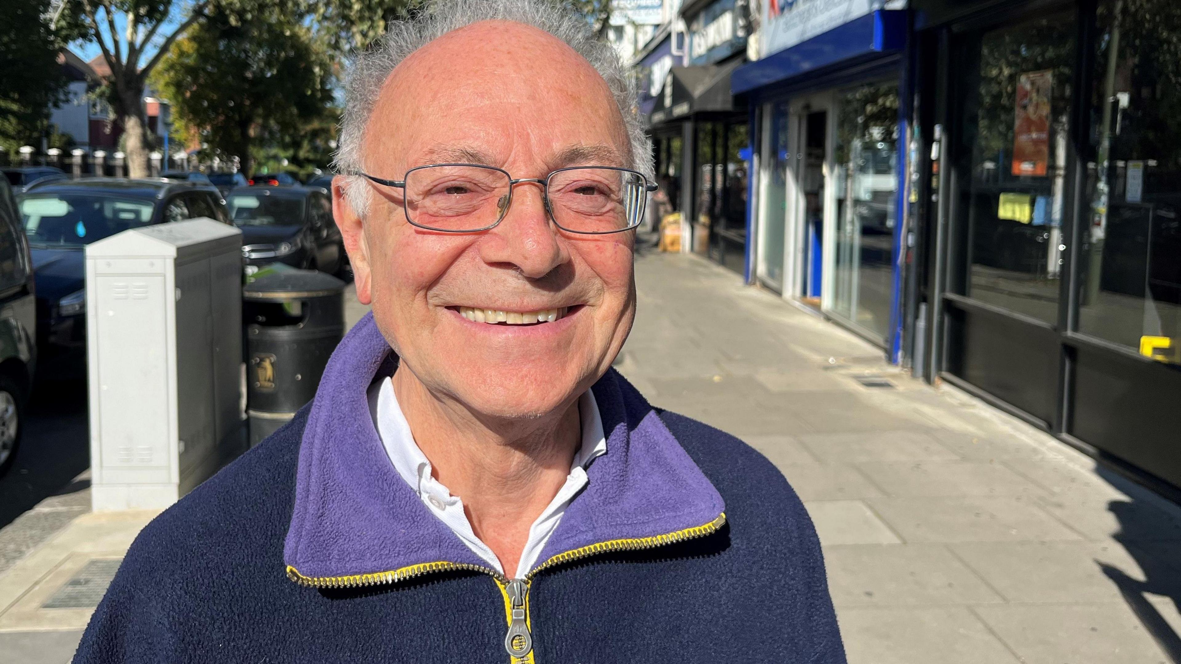 Norman Waidhofer is seen wearing glasses and a fleece jacket smiling on a shopping street, with shop signs and parked cars behind him.