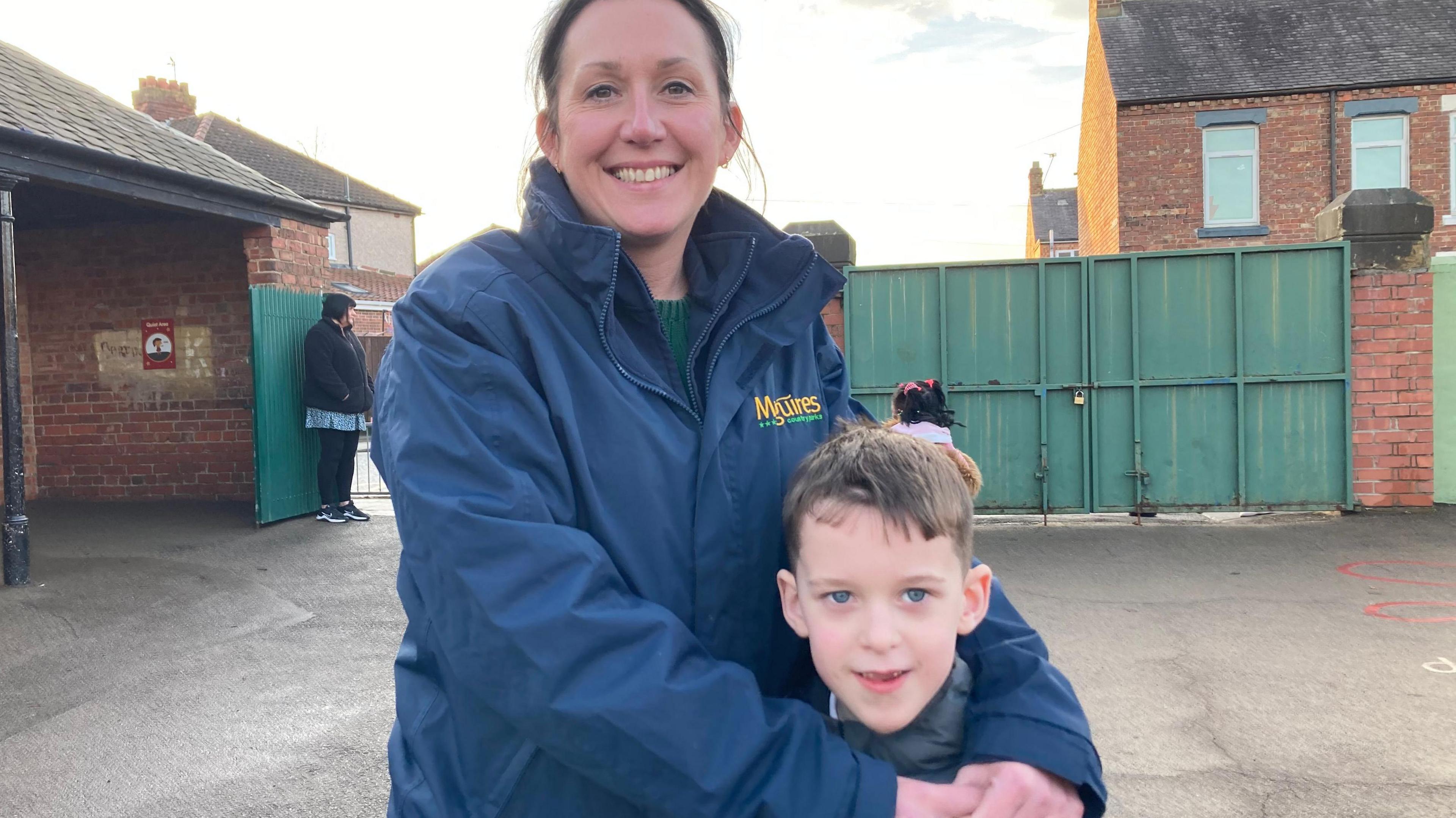 A mother and son stand together smiling in the school playground.