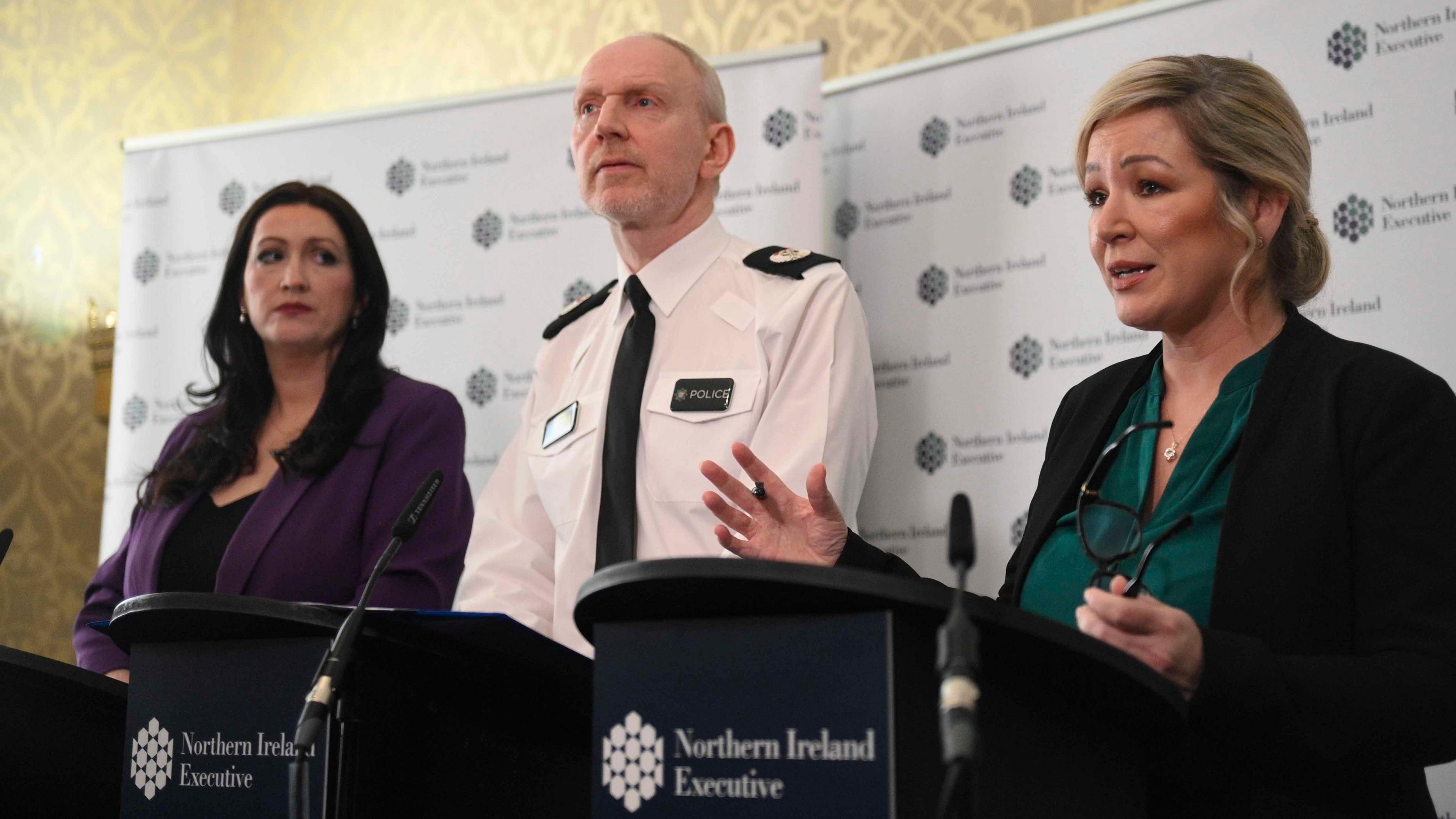 Deputy First Minister Emma Little-Pengelly in a purple suit jacket and black top, Ass Ch Cons Davy Beck in PSNI uniform, white shirt with police badges and black tie, and First Minister Michelle O'Neill wearing a black blazer and green blouse, stood at a NI exuctive backdrop with black podiums and microphones as they held a press conference on Thursday at Stormont